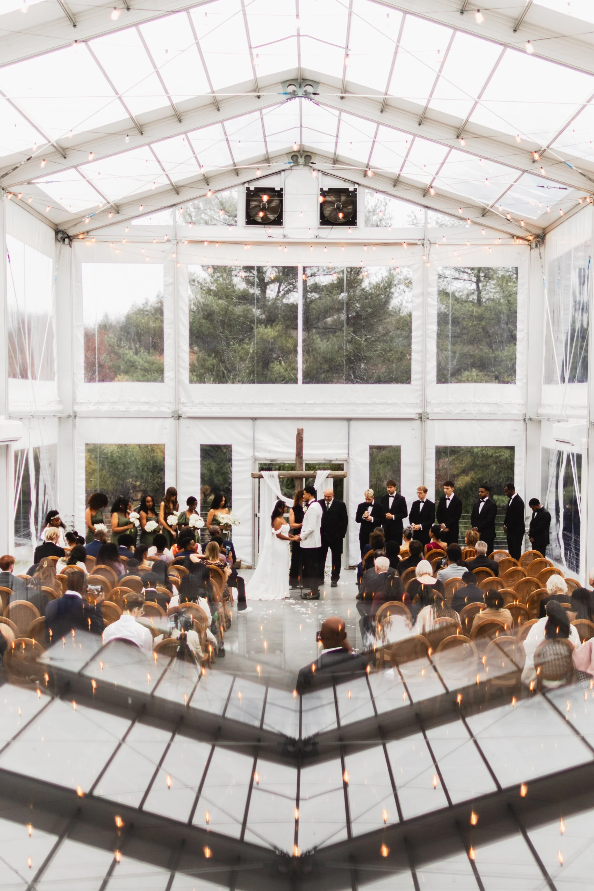 Indoor wedding ceremony under a transparent tent with string lights, featuring a bride and groom exchanging vows in front of a wooden cross, surrounded by bridesmaids and groomsmen, with seated guests watching.