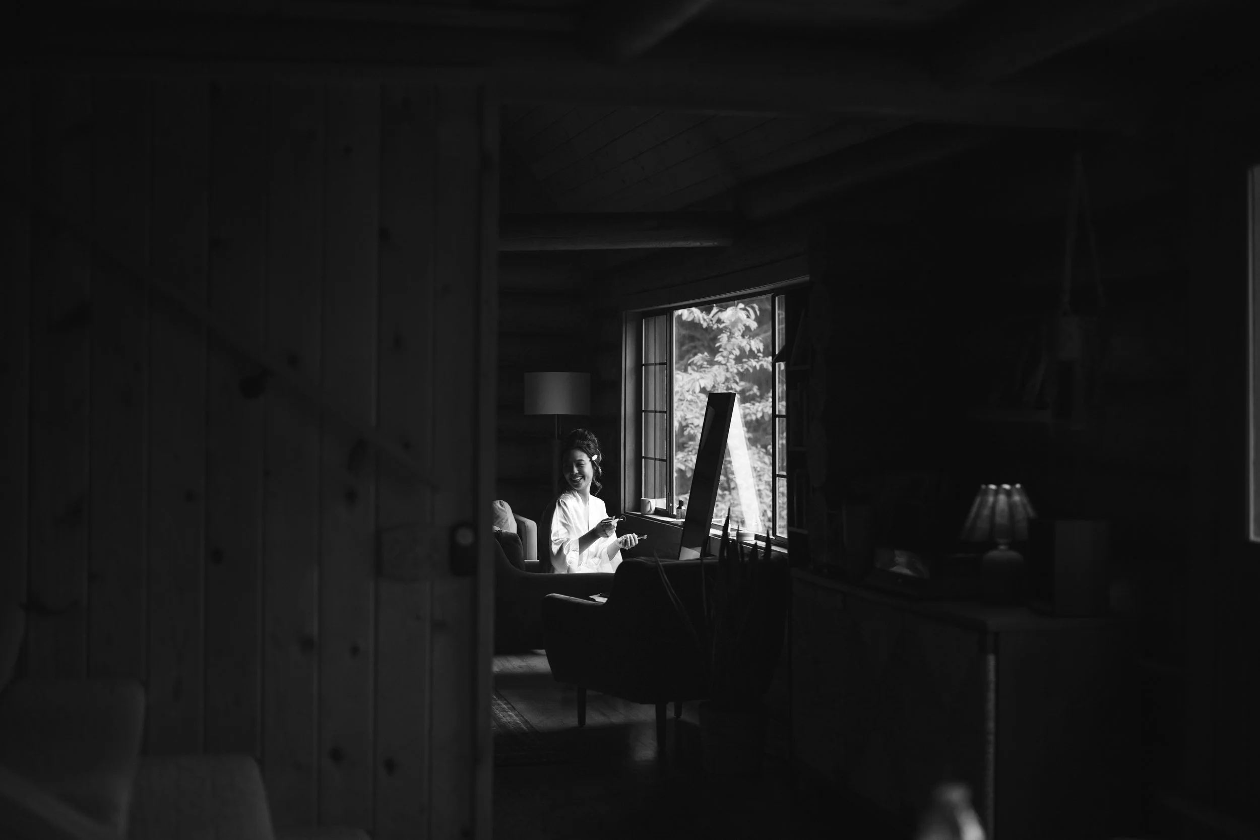 A woman sitting by a window in a cozy wooden interior, smiling and holding a phone, with sunlight streaming in through the open window.