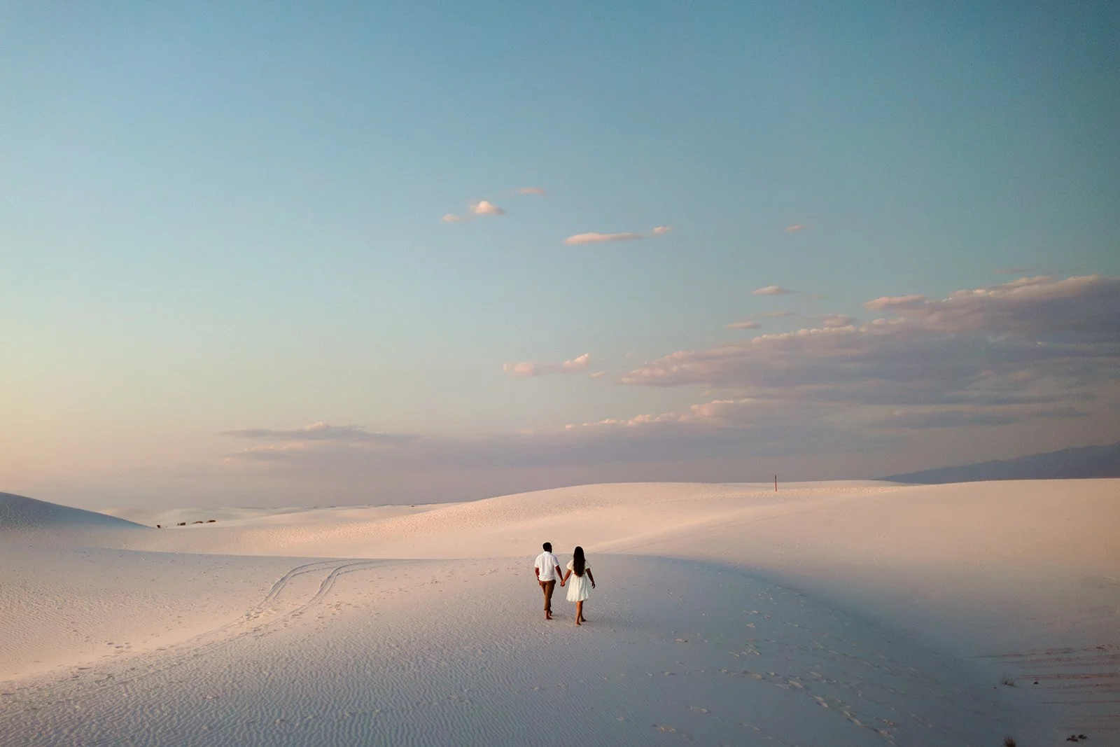 A couple holding hands walking in a vast, white sandy desert landscape under a light blue sky with sparse clouds.
