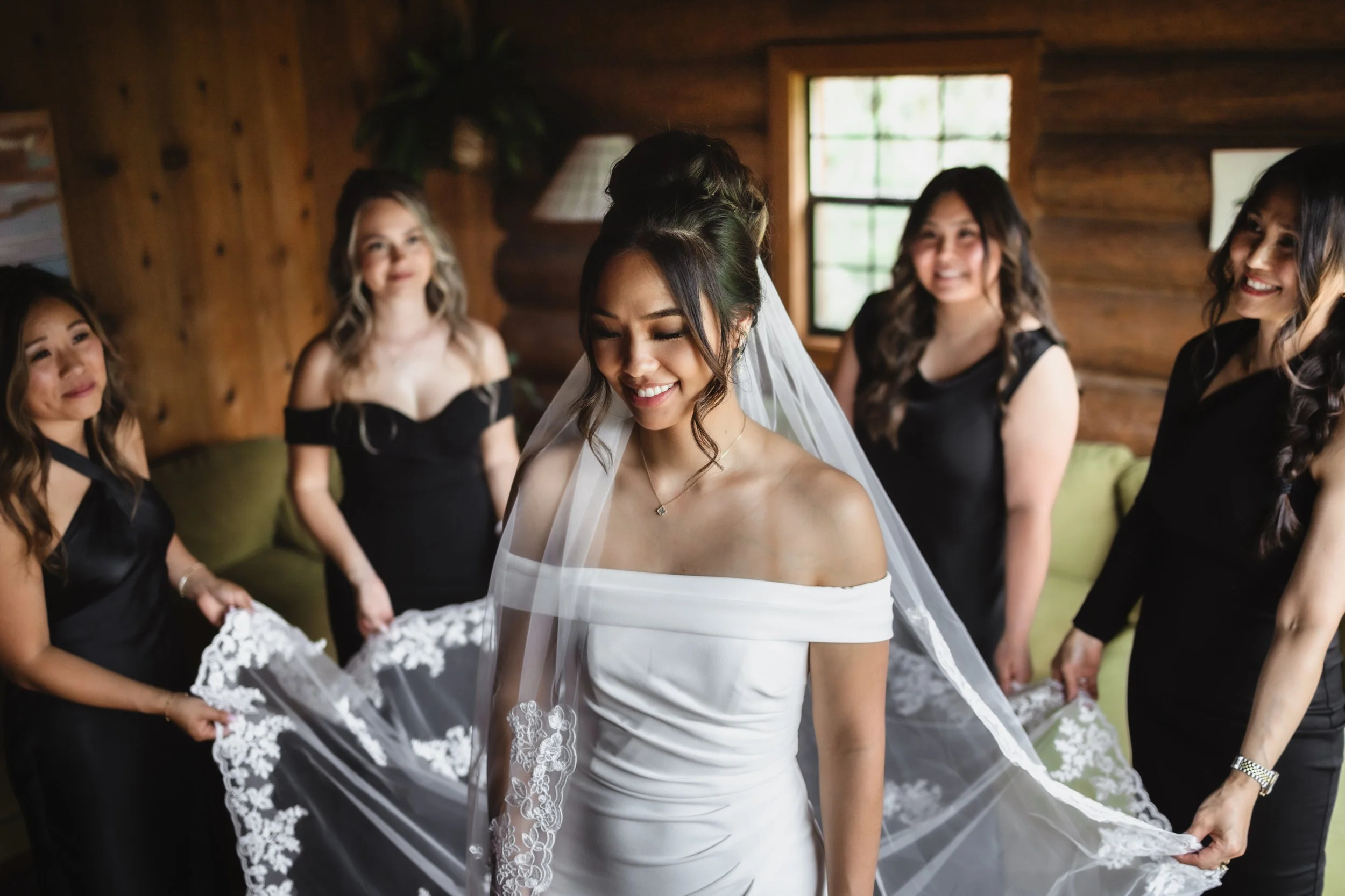 Bride in white off-shoulder wedding dress with veil, smiling with eyes closed, surrounded by four women in black dresses holding her veil, in a wooden cabin interior.