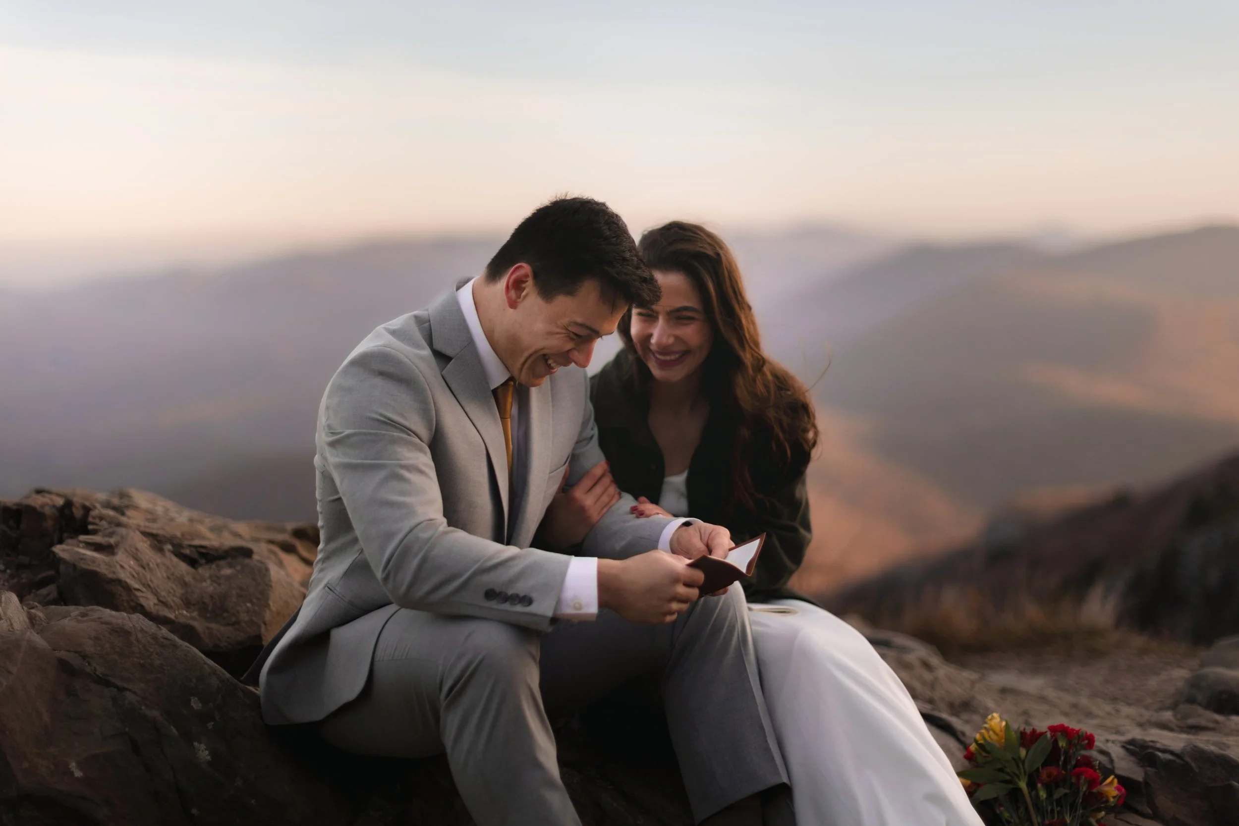 A couple dressed in formal wedding attire sitting on rocks outdoors, smiling and looking at a phone, with a mountain landscape in the background during sunset.