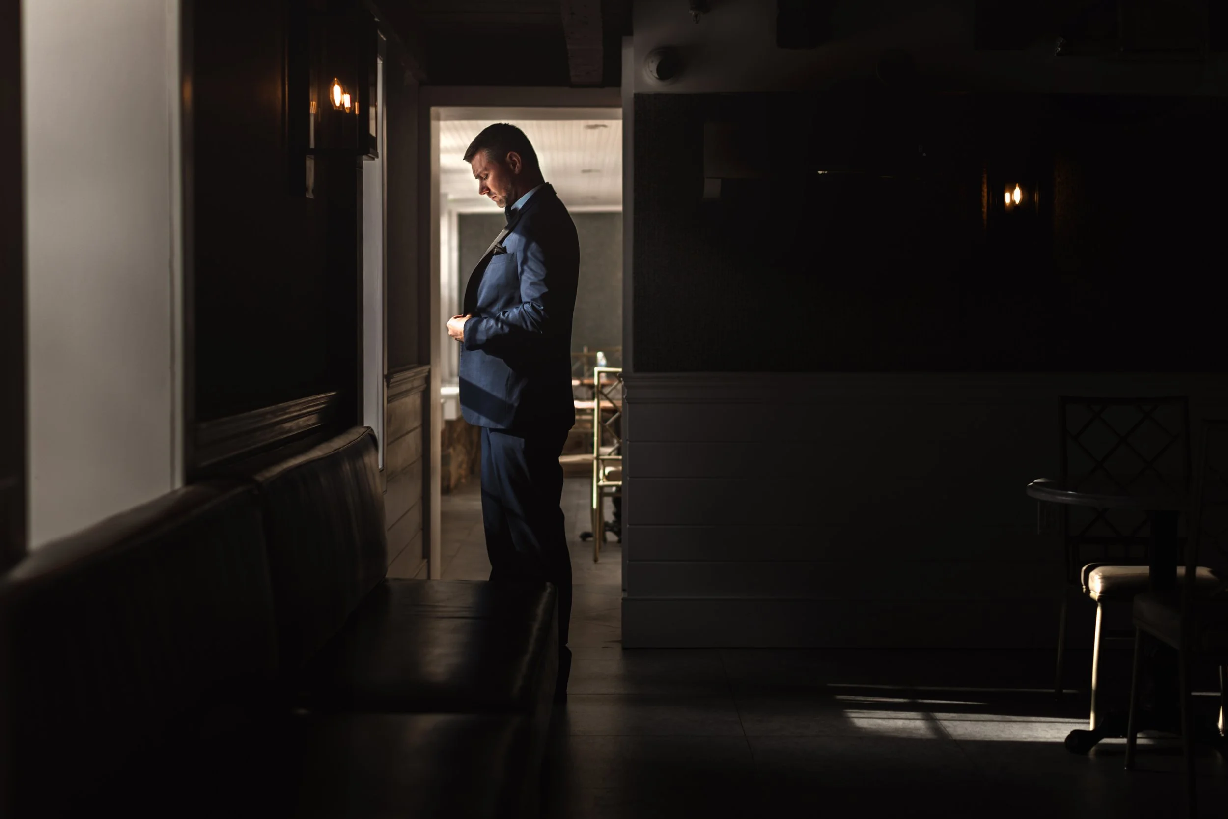 Man in a blue suit standing in a dimly lit room, looking down, with sunlight coming from behind him.