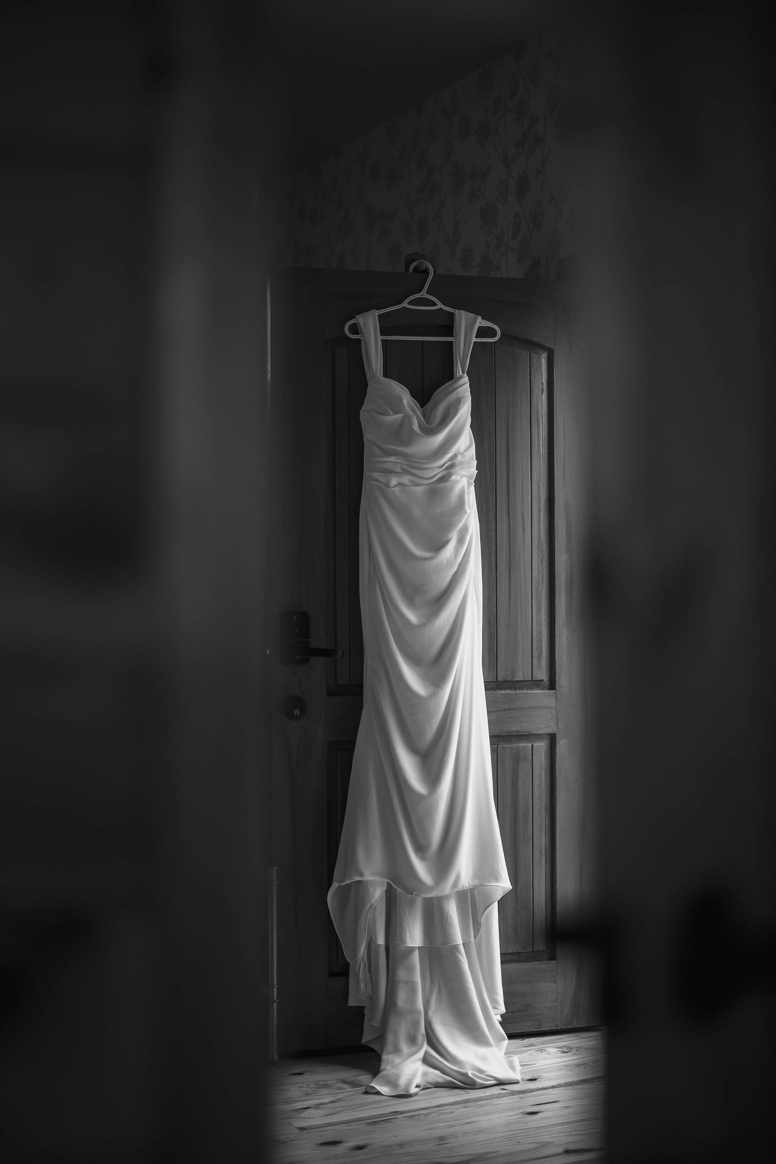 A white wedding dress hanging on a hanger on a wooden door, captured through a partially opened door in black and white.