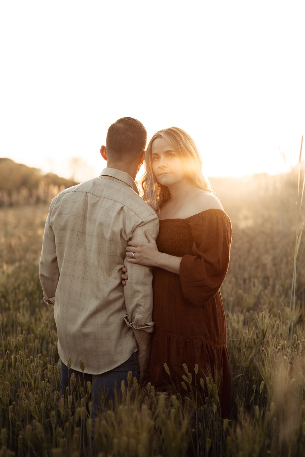 A man and a woman standing close in a field of grass during sunset, with the man facing away and the woman looking at the camera.