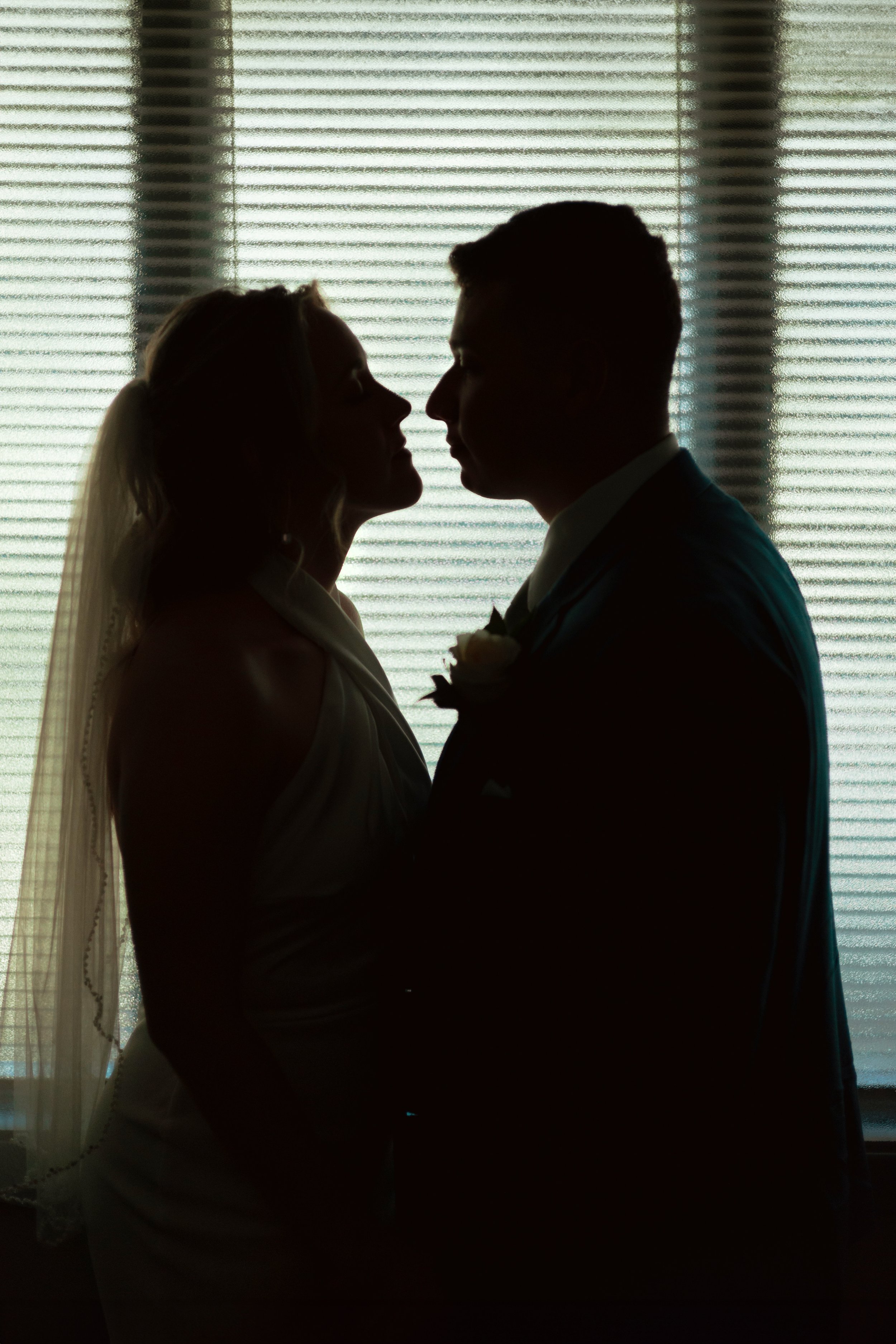 Silhouettes of a bride and groom facing each other during a wedding, with a window and blinds in the background.