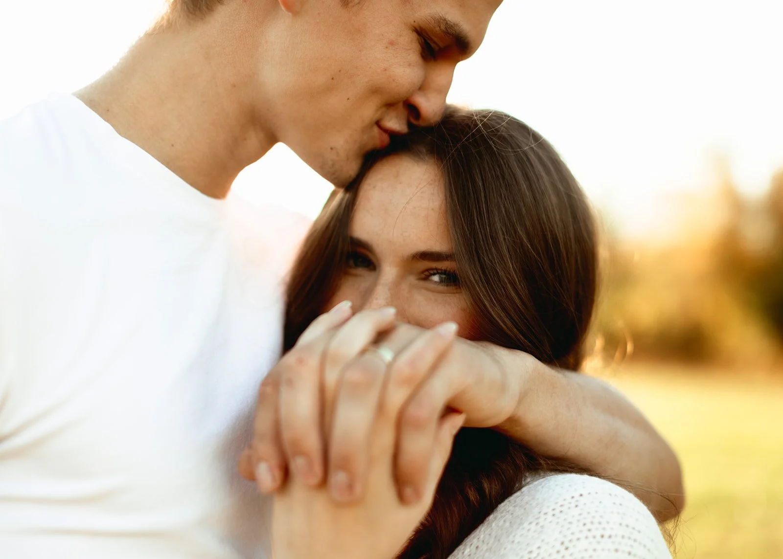 A young man kisses a young woman on the head while she smiles and looks at the camera, in an outdoor setting during sunset.
