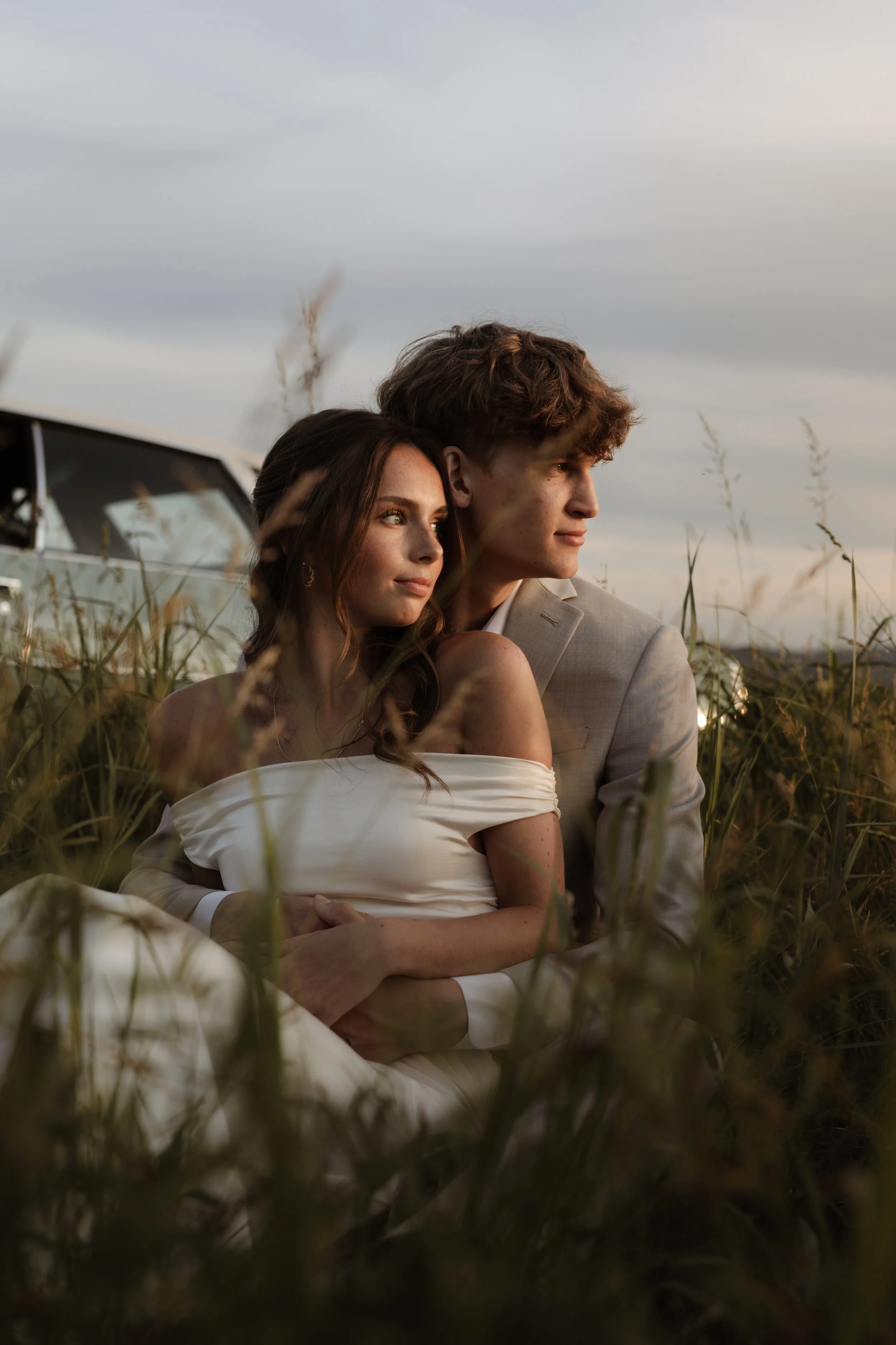 A couple in wedding attire sitting close together in a grassy field at sunset, with a vehicle in the background.