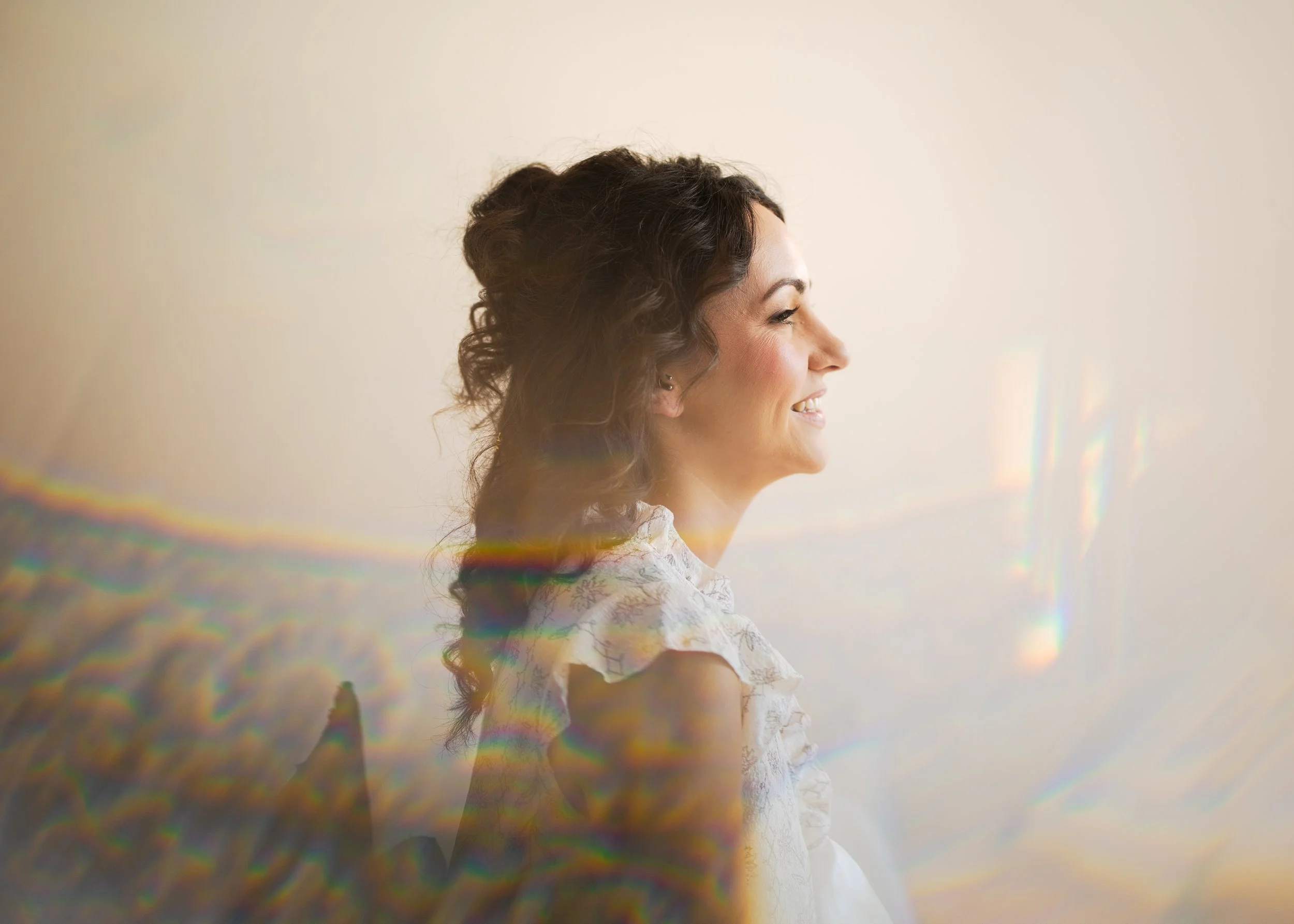 A woman with dark, curly hair smiling, sitting in front of a light background, with rainbow light effects across her and the scene.
