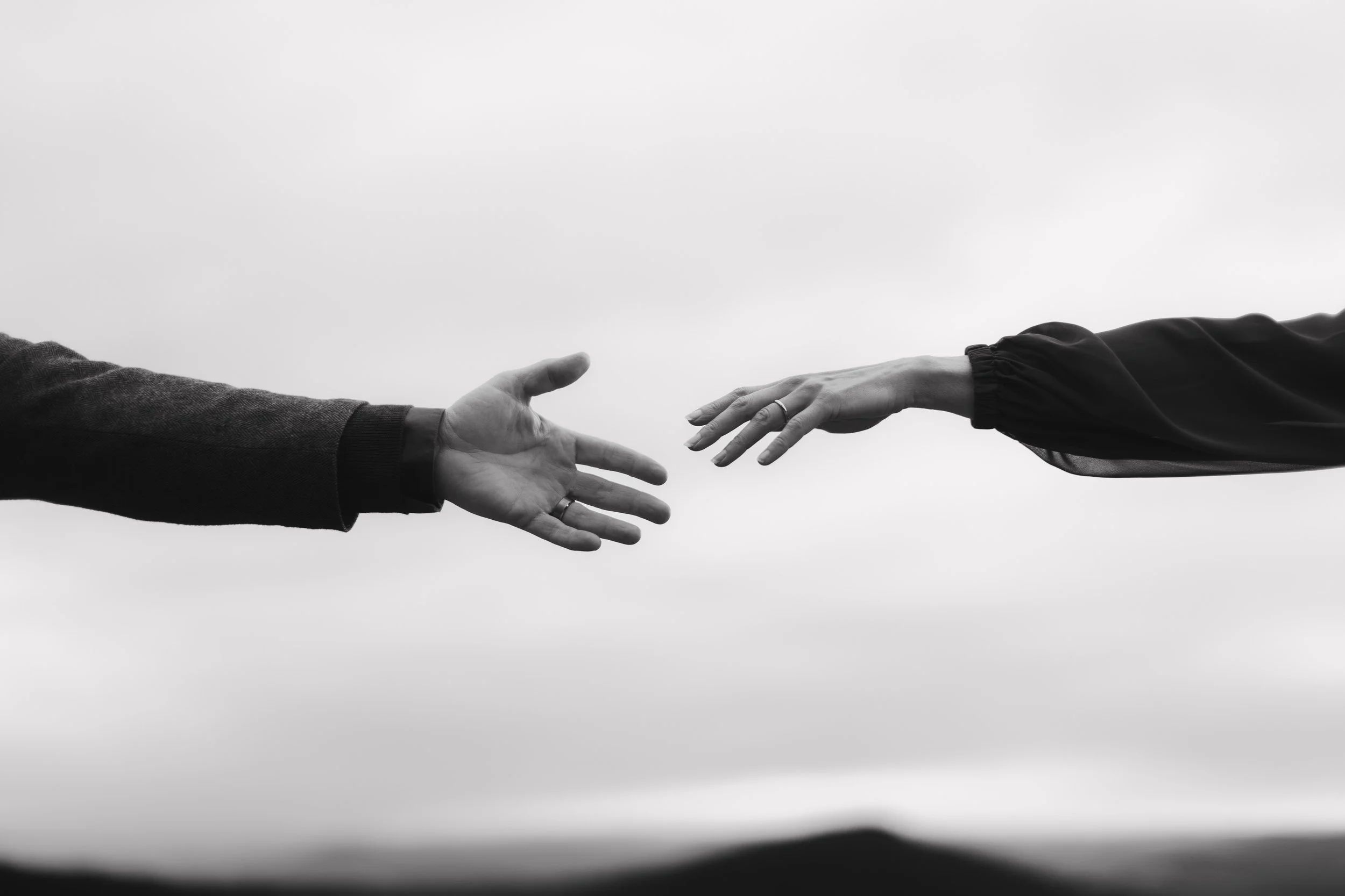 Two people reaching out to each other with their hands, the person on the left is wearing a dark sweater and the person on the right is wearing a long sleeve top and a ring.