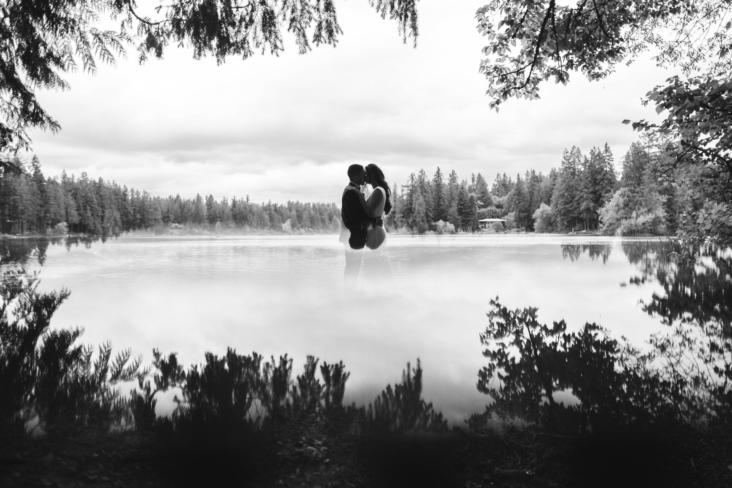 A couple standing close together, embracing and kissing by a lake with trees in the background, in black and white.