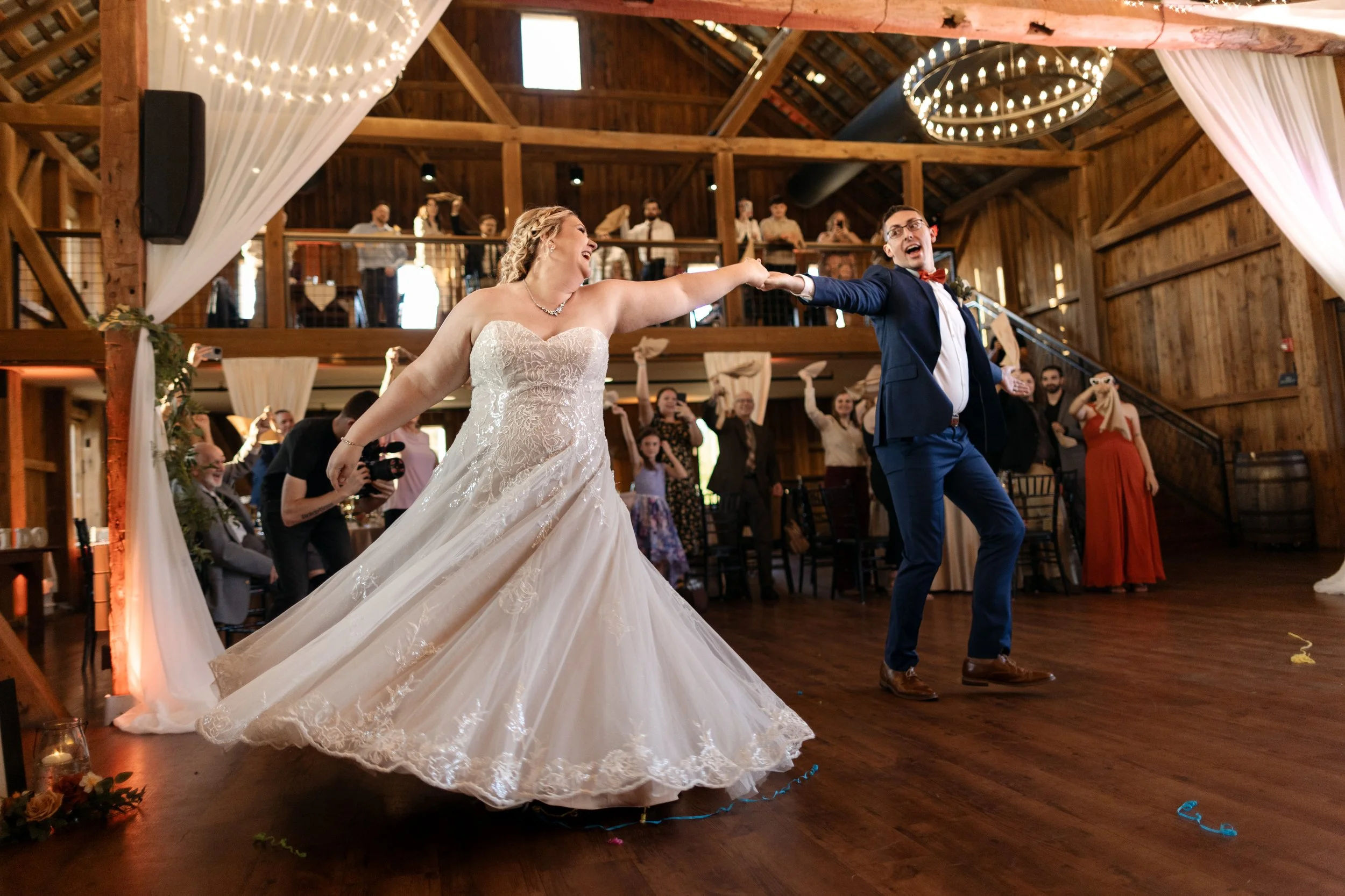 A bride and groom are dancing at their wedding reception in a rustic barn, holding hands and smiling, with guests in the background cheering.
