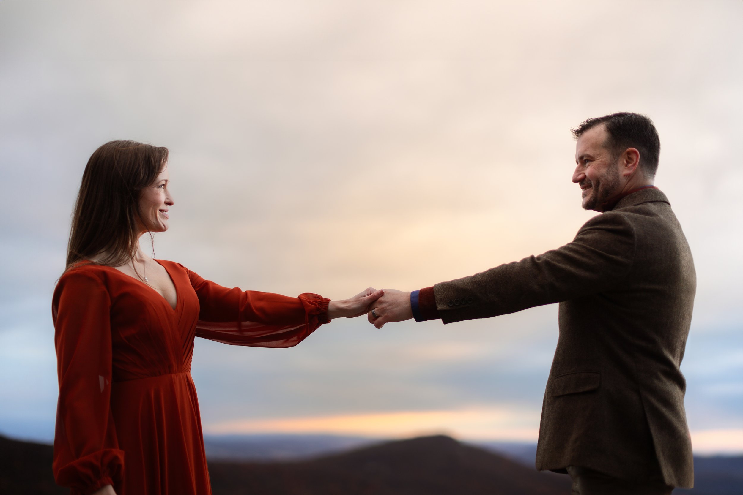 A man and woman holding hands, smiling at each other outdoors at sunset, with a landscape background.