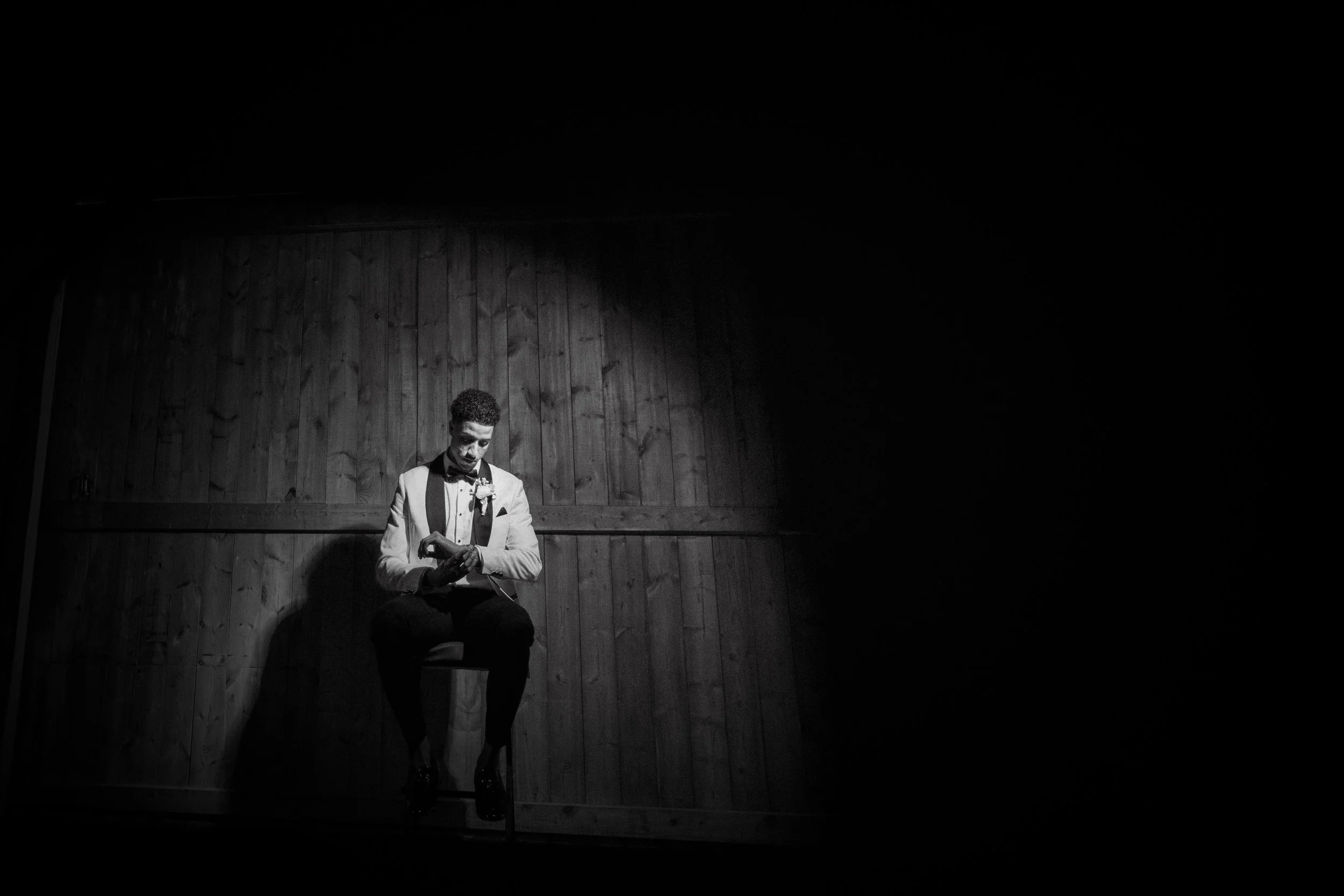 A man in a tuxedo sits on a chair in a dimly lit wooden room, looking at his watch.