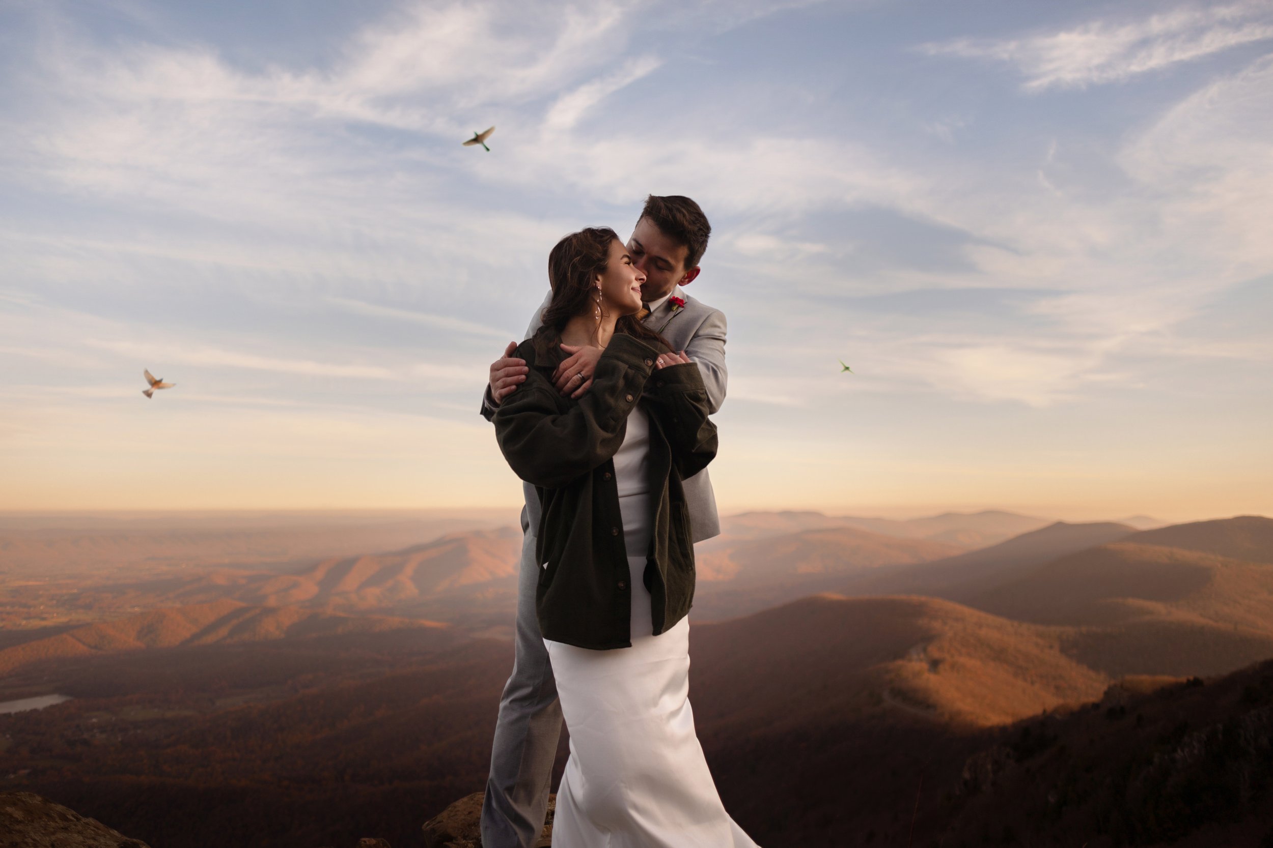A young couple embraces on a mountain top during sunset, with a scenic view of rolling hills and a sky with clouds and flying birds in the background.