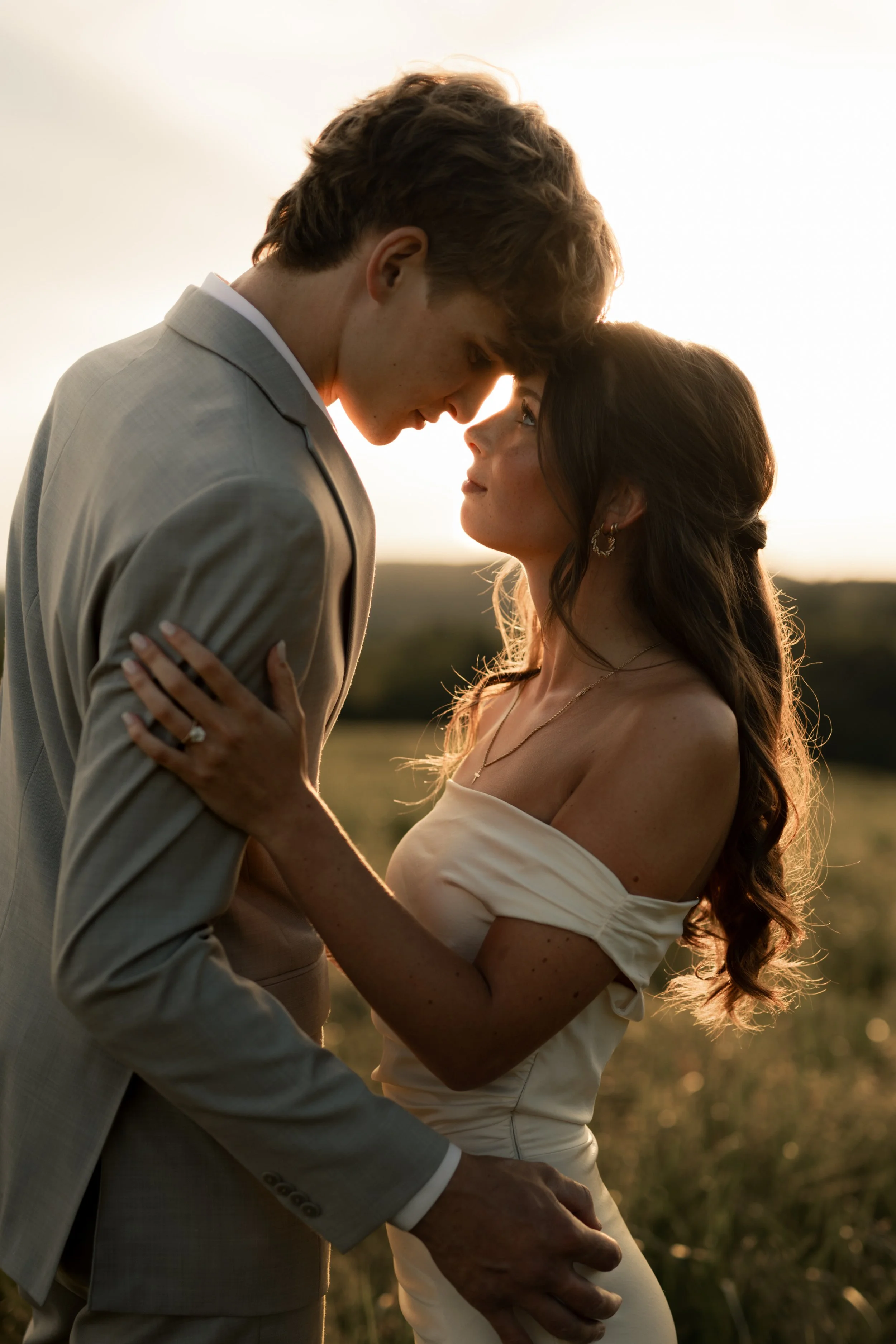 A couple in a romantic embrace in a field at sunset, touching foreheads and gazing into each other's eyes.