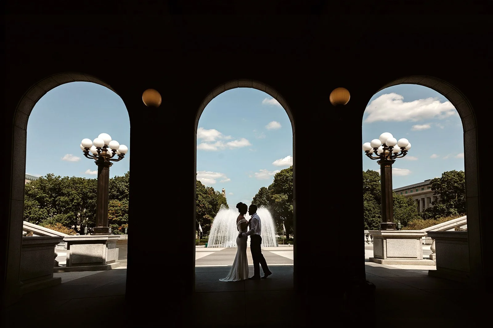 Silhouetted couple in wedding attire standing close together under an archway, with fountains and trees in the background, during daytime.