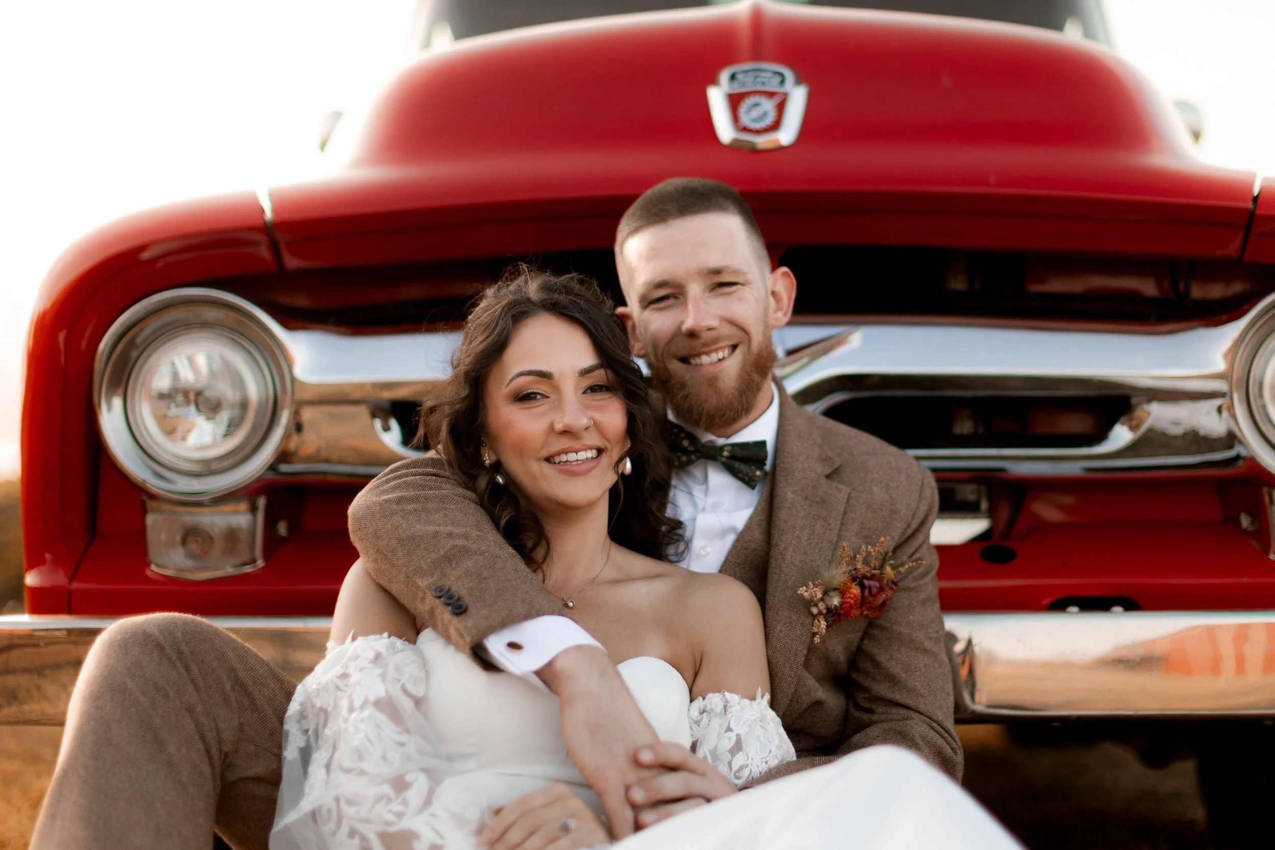 A happy couple in wedding attire sitting in front of a vintage red car, smiling at the camera.