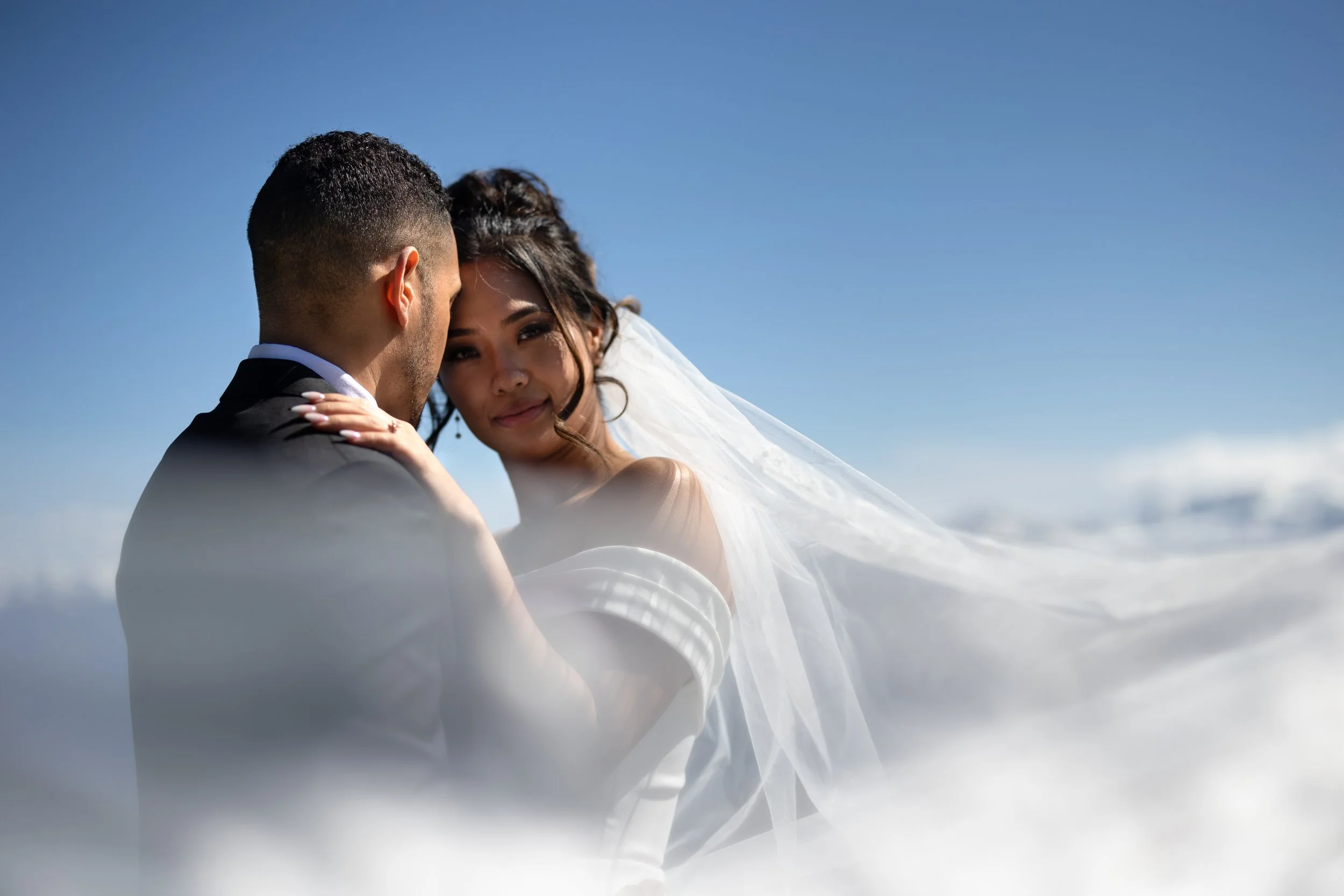 A couple in wedding attire standing close, with the woman's head resting on the man's shoulder, against a blue sky and clouds.