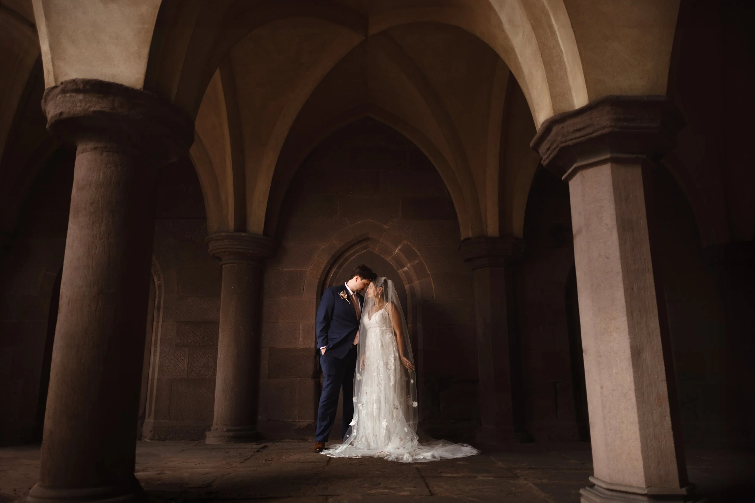 Black and white photo of a bride in a wedding dress with a veil holding a bouquet, standing in a hallway with a man in a suit blurred in the background.
