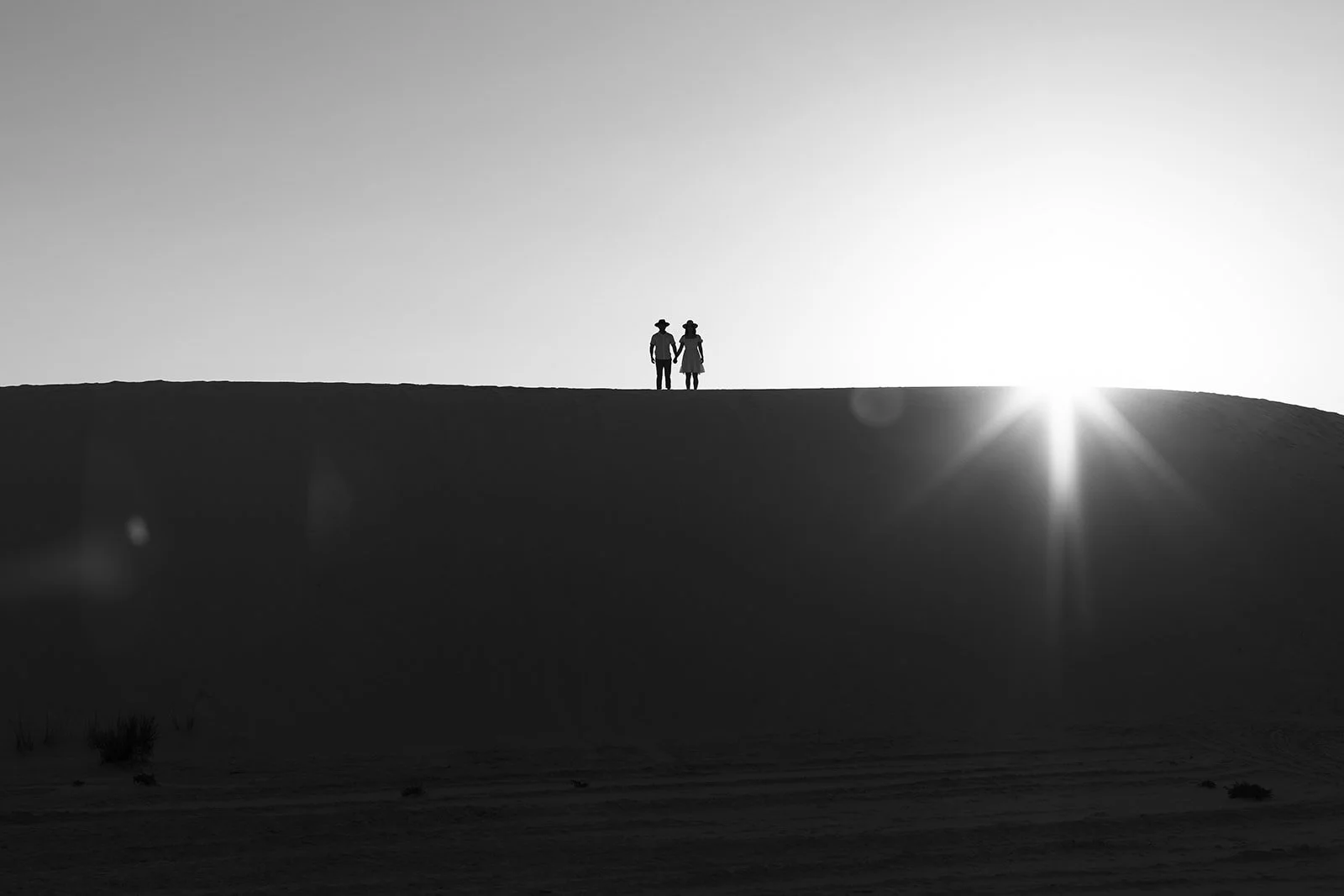 Silhouette of a couple holding hands standing on a hill at sunset with the sun near the horizon and sunbeams radiating out.