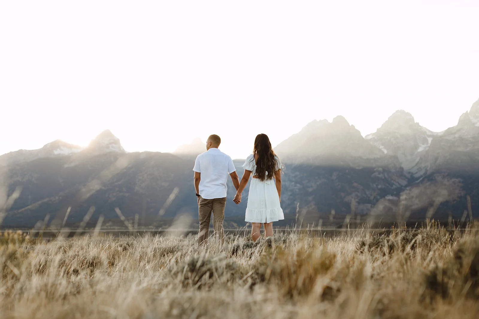 A couple holding hands walking through a field with mountains in the background at sunset.