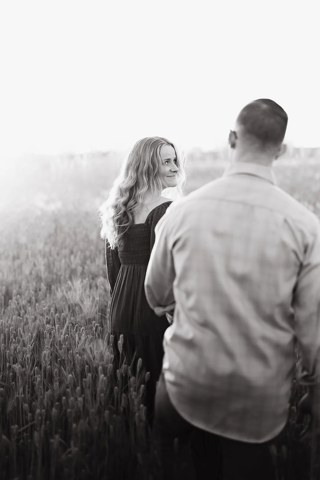A woman with long, wavy hair in a black dress standing in a field of tall grass, smiling and looking at a man in a light-colored shirt with his back to the camera, outdoors during daylight.