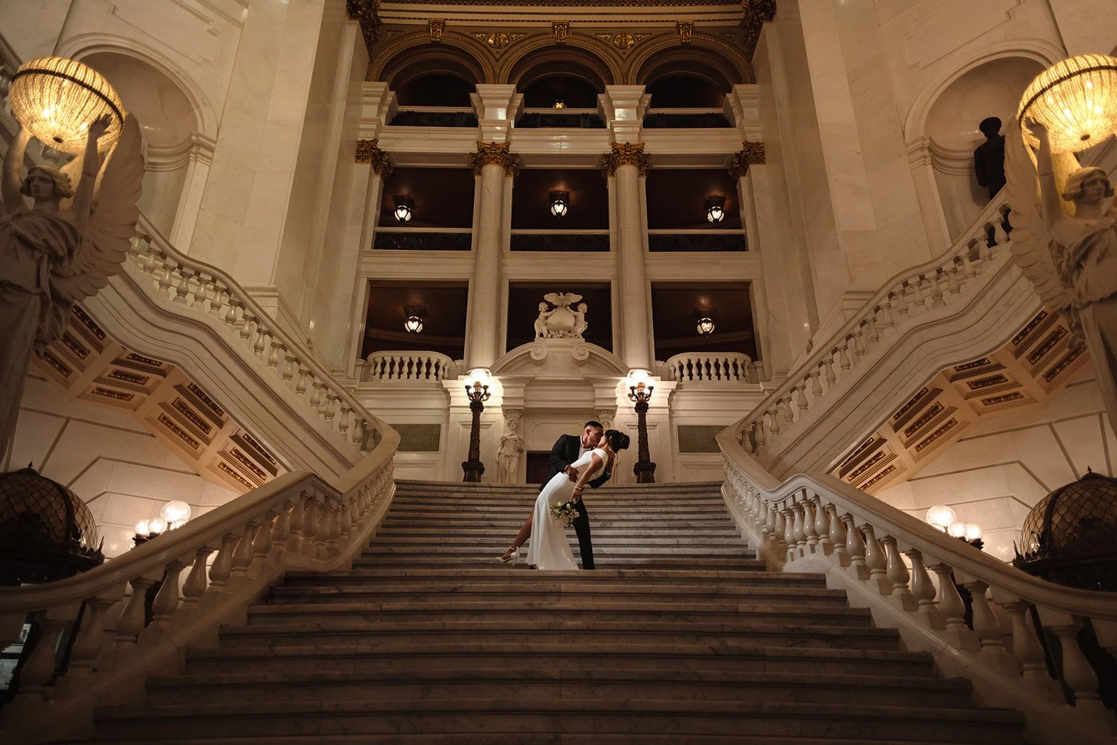 A bride and groom kissing on grand marble staircase inside an elegant, ornate building with marble columns, sculptures, and warm lighting.