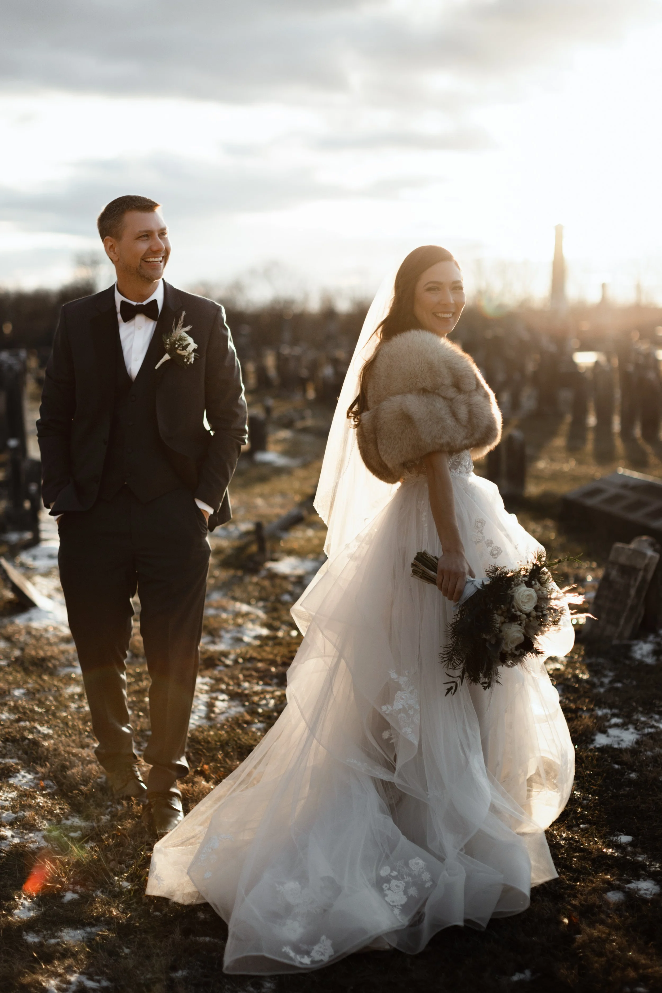 A bride and groom standing in a cemetery during sunset, with the bride holding a bouquet and wearing a fur shawl over her wedding dress, and the groom in a black tuxedo with a boutonniere, both smiling.