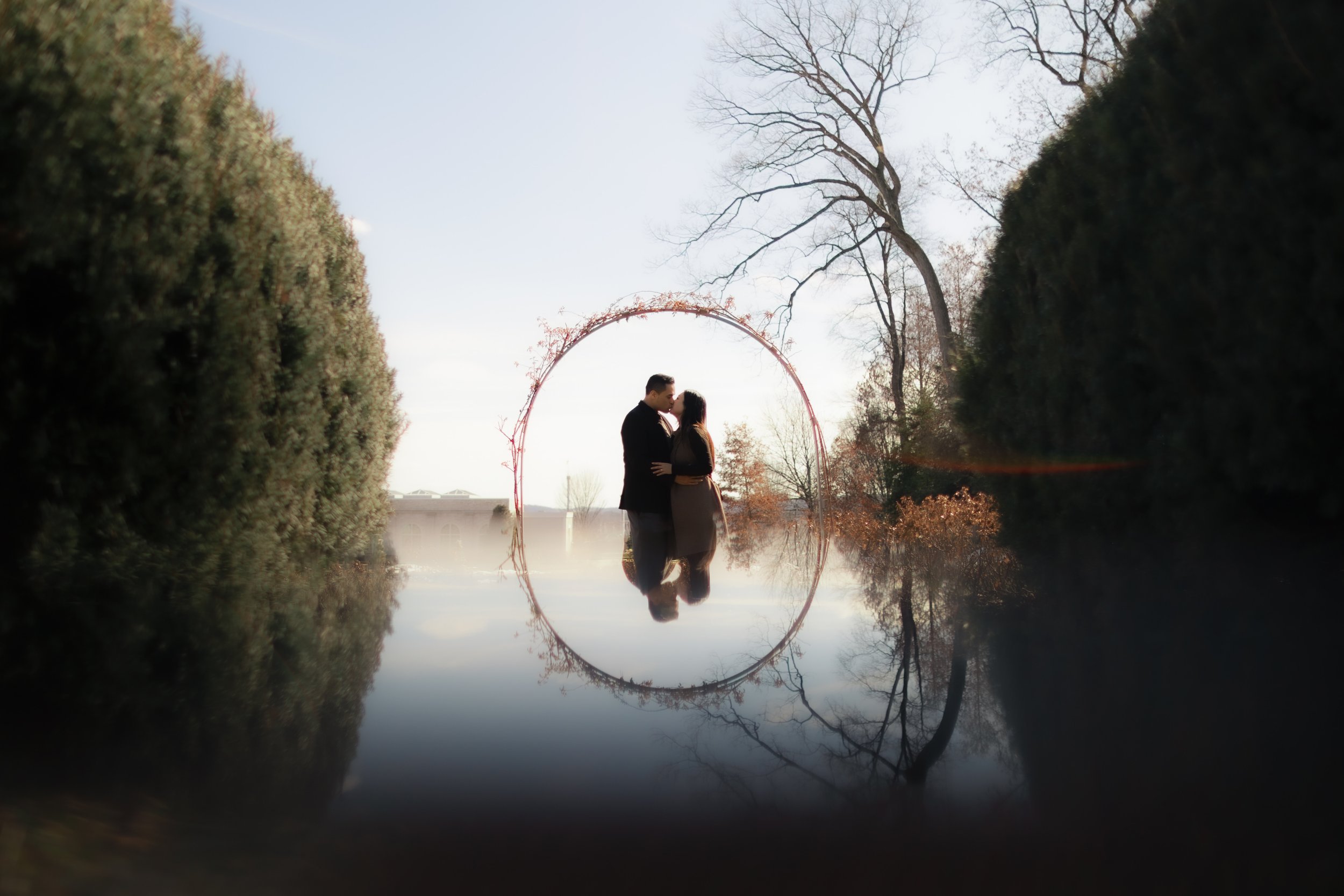 A couple kissing under a decorated arch on a foggy day with trees and water reflection in the background.
