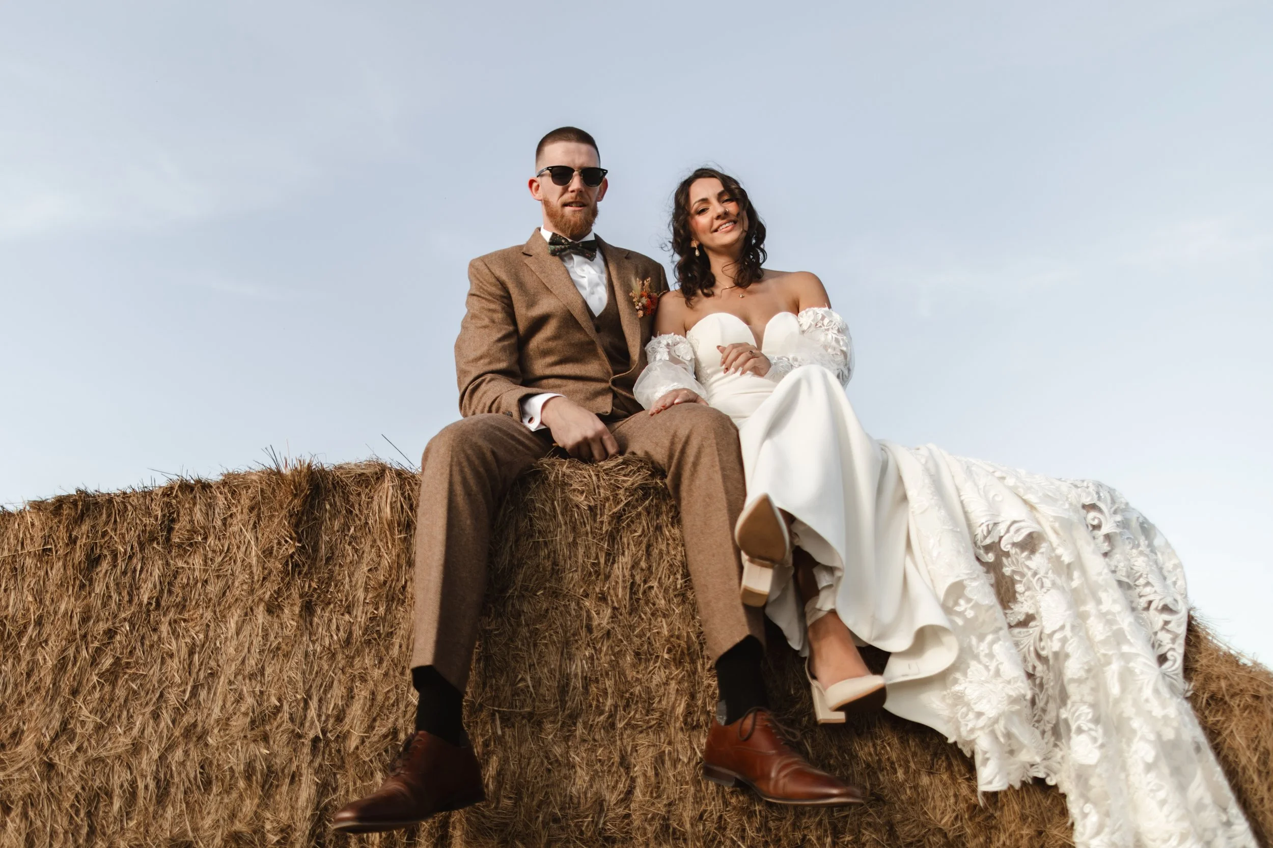 A man and woman sitting on a hay bale outdoors, the man in a brown suit and sunglasses, the woman in a white wedding dress, both smiling.