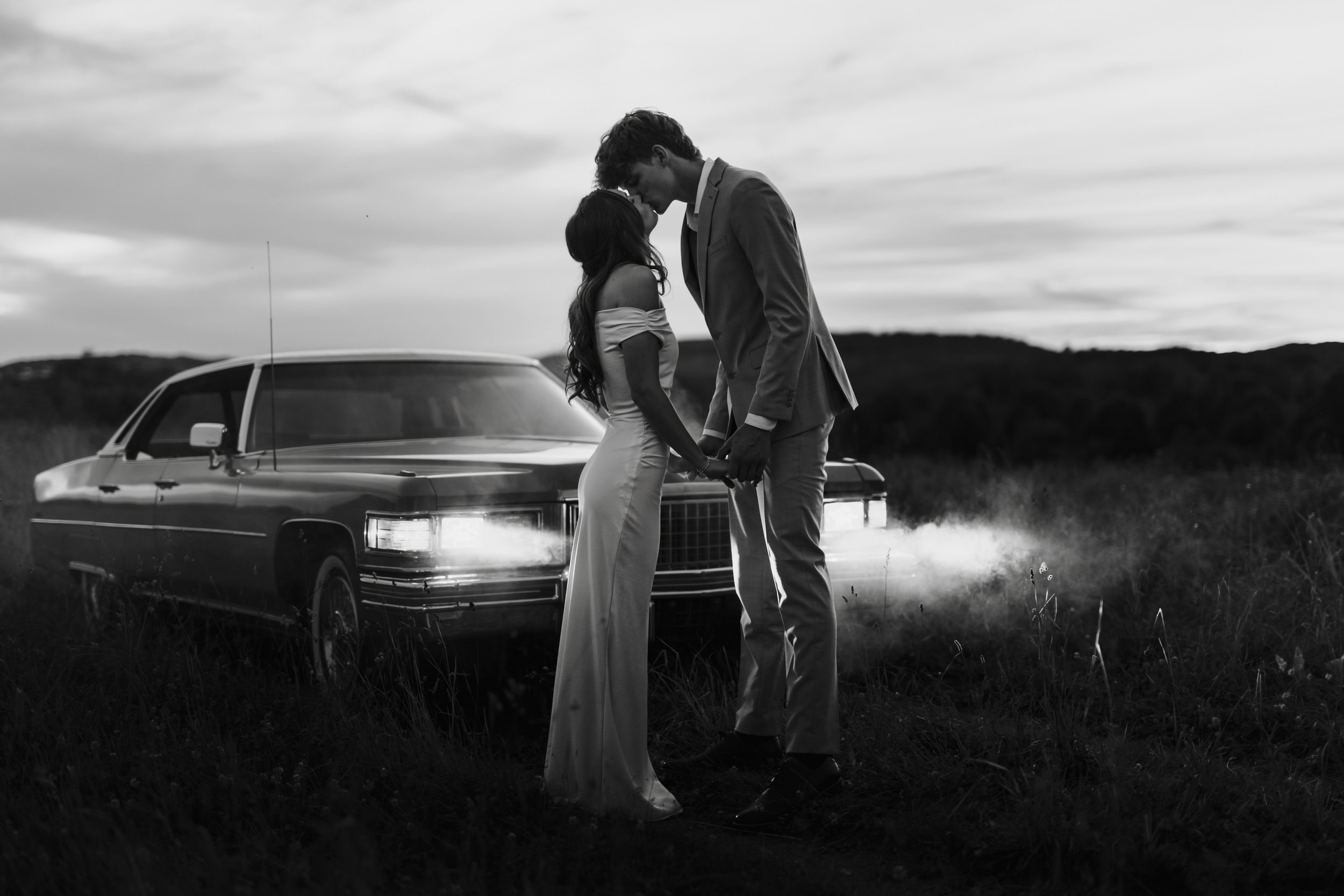 A couple dressed in wedding attire holding hands in front of a vintage car on a grassy field during dusk, black and white photograph
