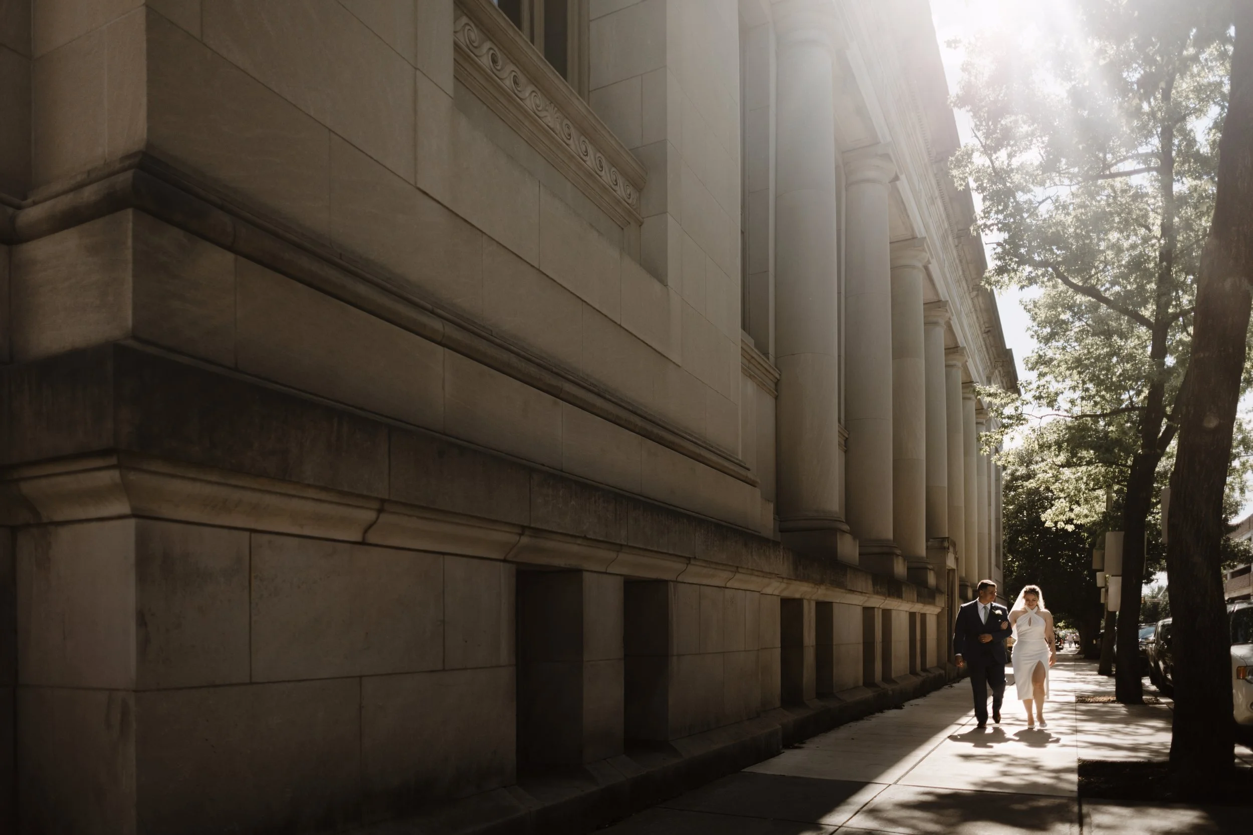 A couple dressed in formal attire walking on a sidewalk next to a large stone building with tall columns, sunlight streaming through the trees.