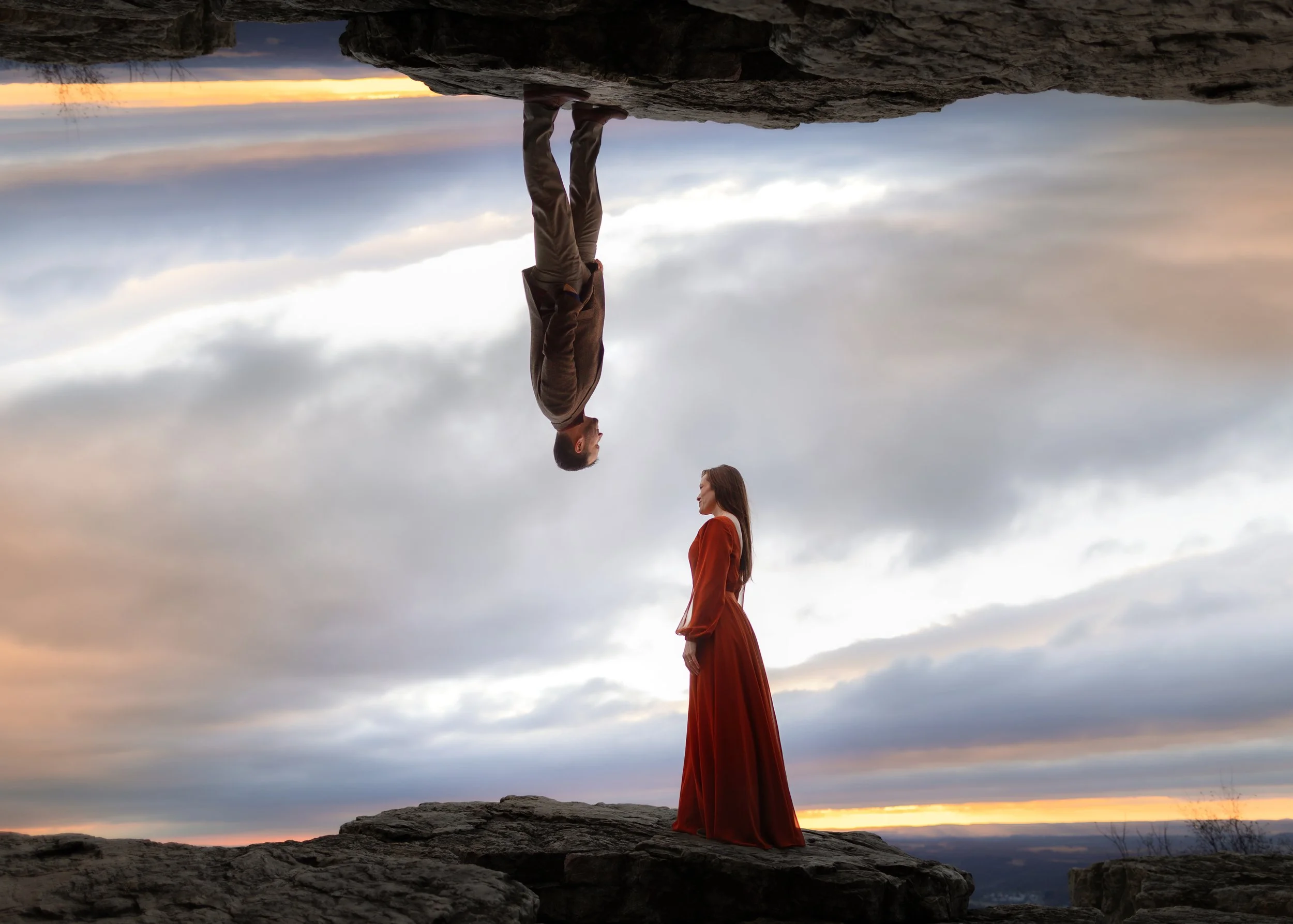 A man and a woman face each other on a rocky landscape at sunset, with the man standing on a rock below and the woman standing on a higher rock. The sky is cloudy with a horizon glow.