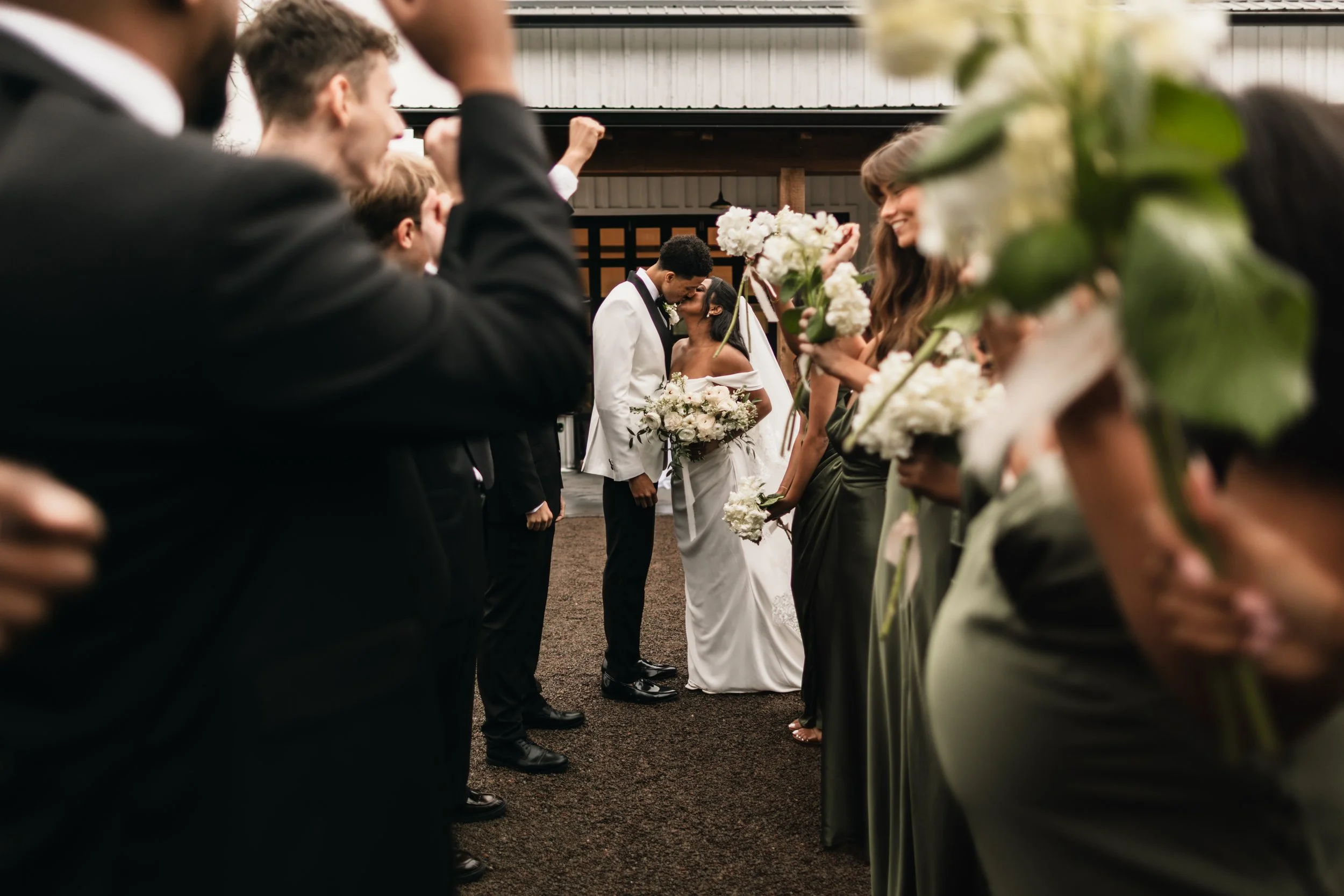 A bride and groom share a kiss at their wedding surrounded by friends and family who are holding bouquets of white flowers, standing in two lines.