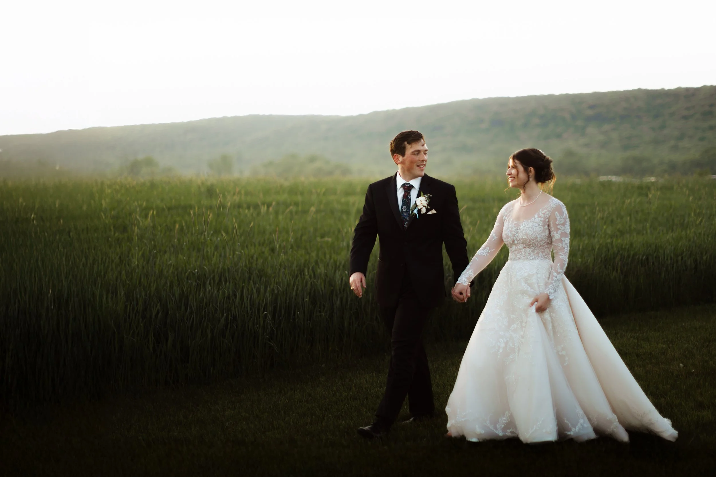 Bride and groom walking through a field at sunset, showcasing stunning sun flare and rustic elegance. Dramatic golden hour wedding photos taken by a professional photographer in Hamburg, Berks County, PA.