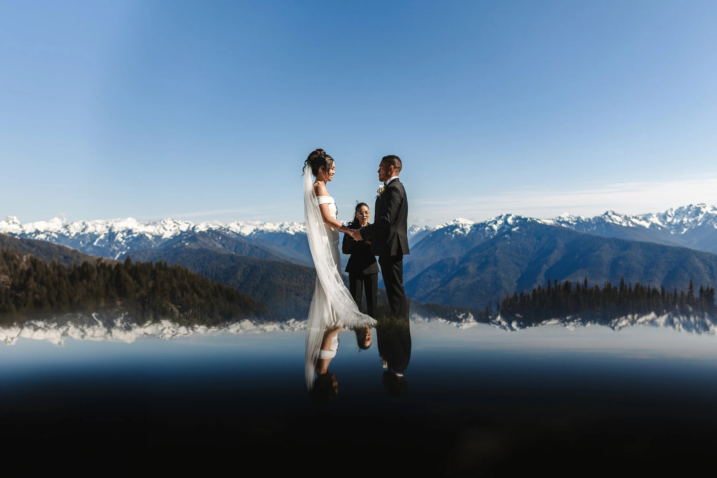 A bride and groom exchanging vows during their outdoor wedding ceremony, with snow-capped mountains and a clear blue sky in the background, reflected in the water below.