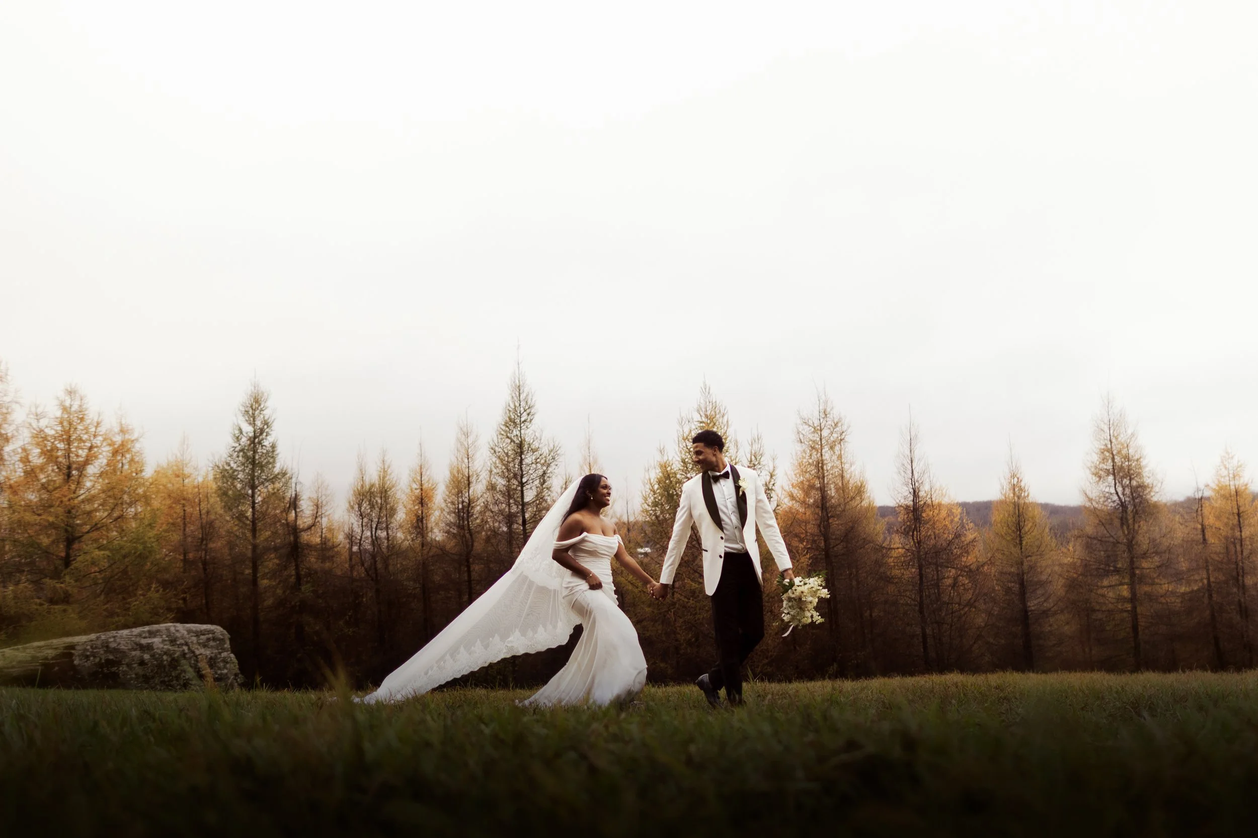 A bride and groom in wedding attire holding hands and walking across a grassy field with trees in fall colors in the background.