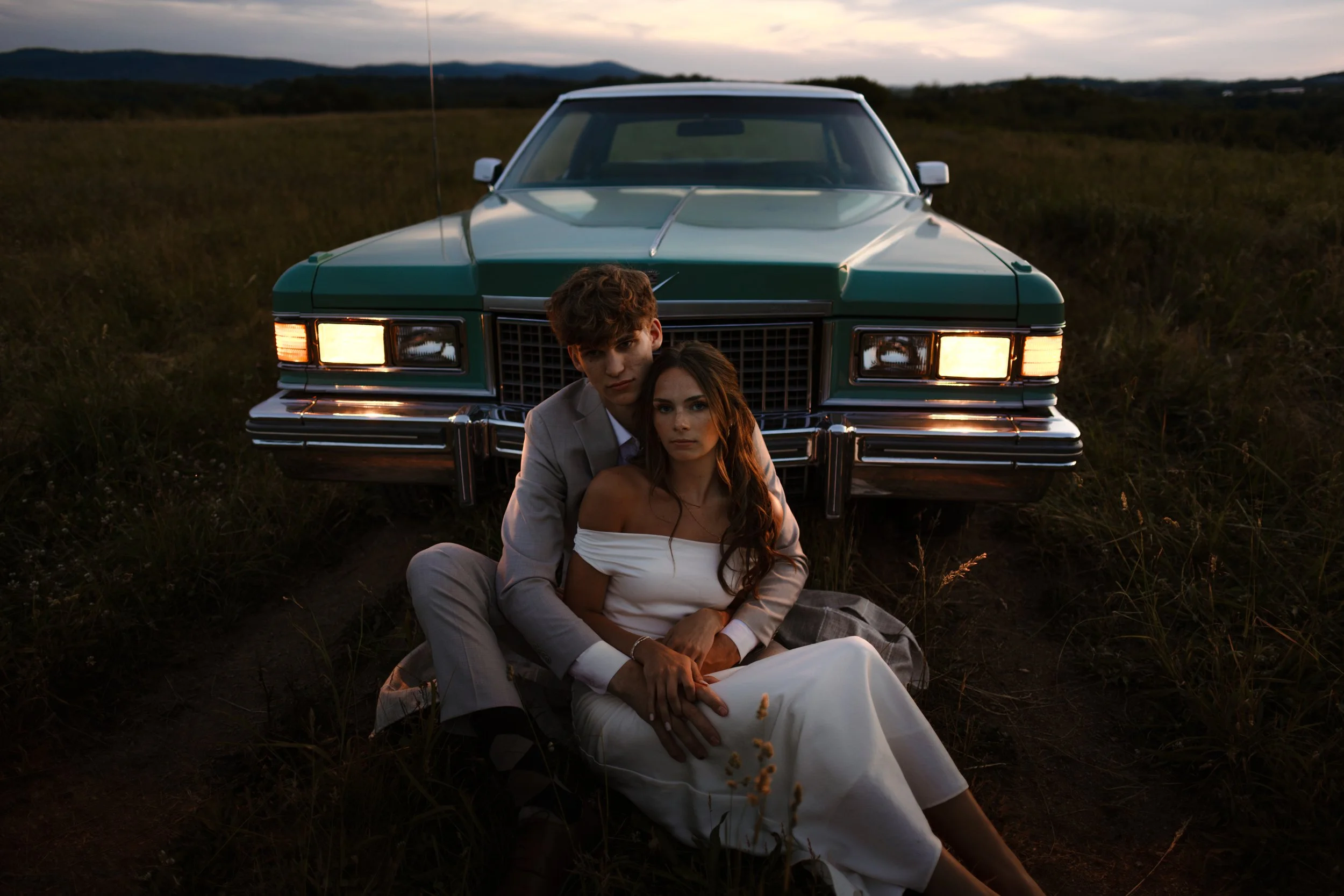 A young couple in formal attire sitting on the ground in front of a green vintage car during sunset in an open field.