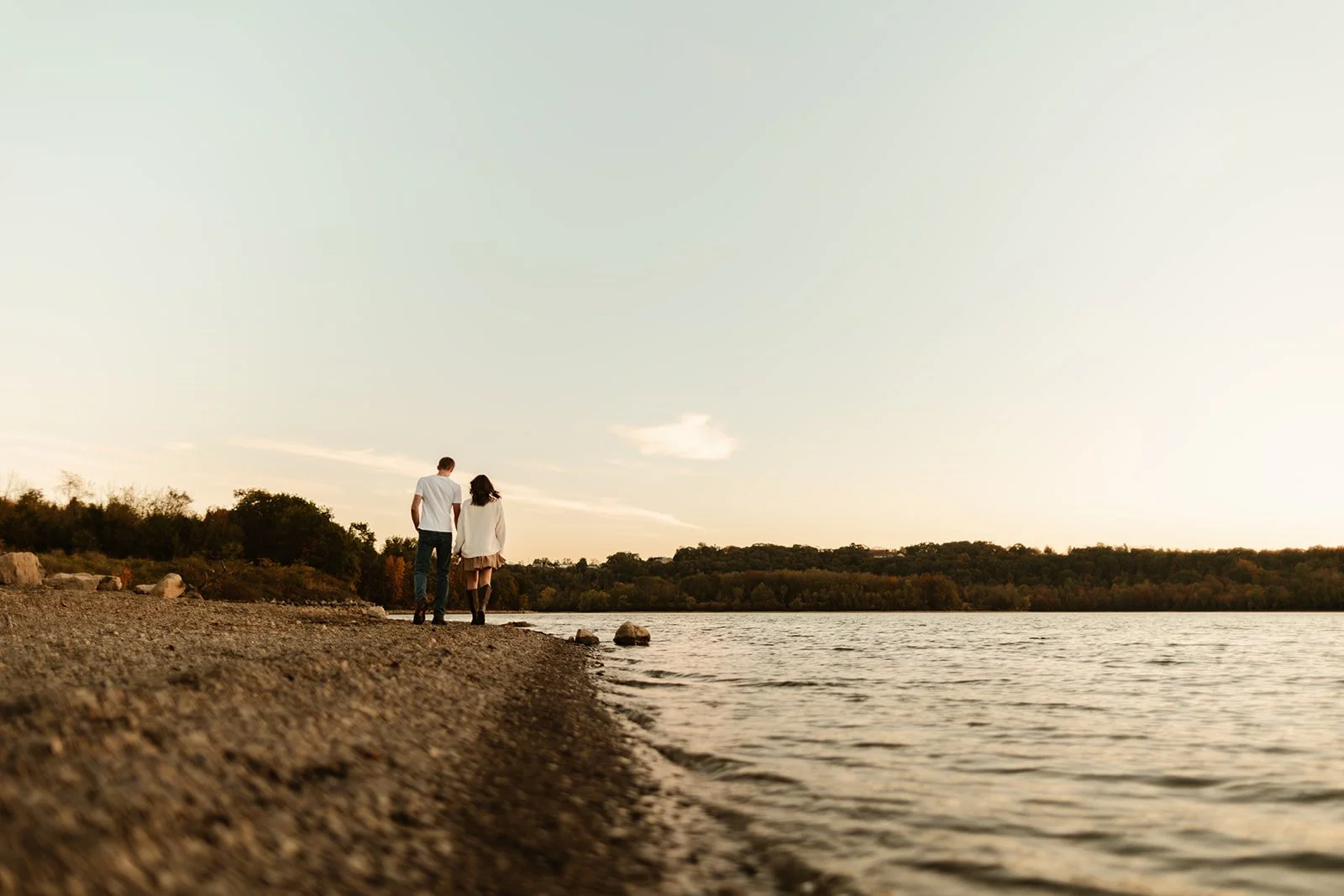 A couple walking along a shoreline during sunset near a body of water with trees in the background.