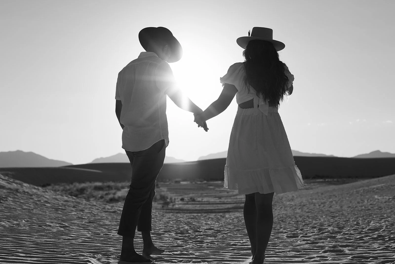 A black and white photo of a couple holding hands while walking in a desert landscape during sunset, both wearing hats.