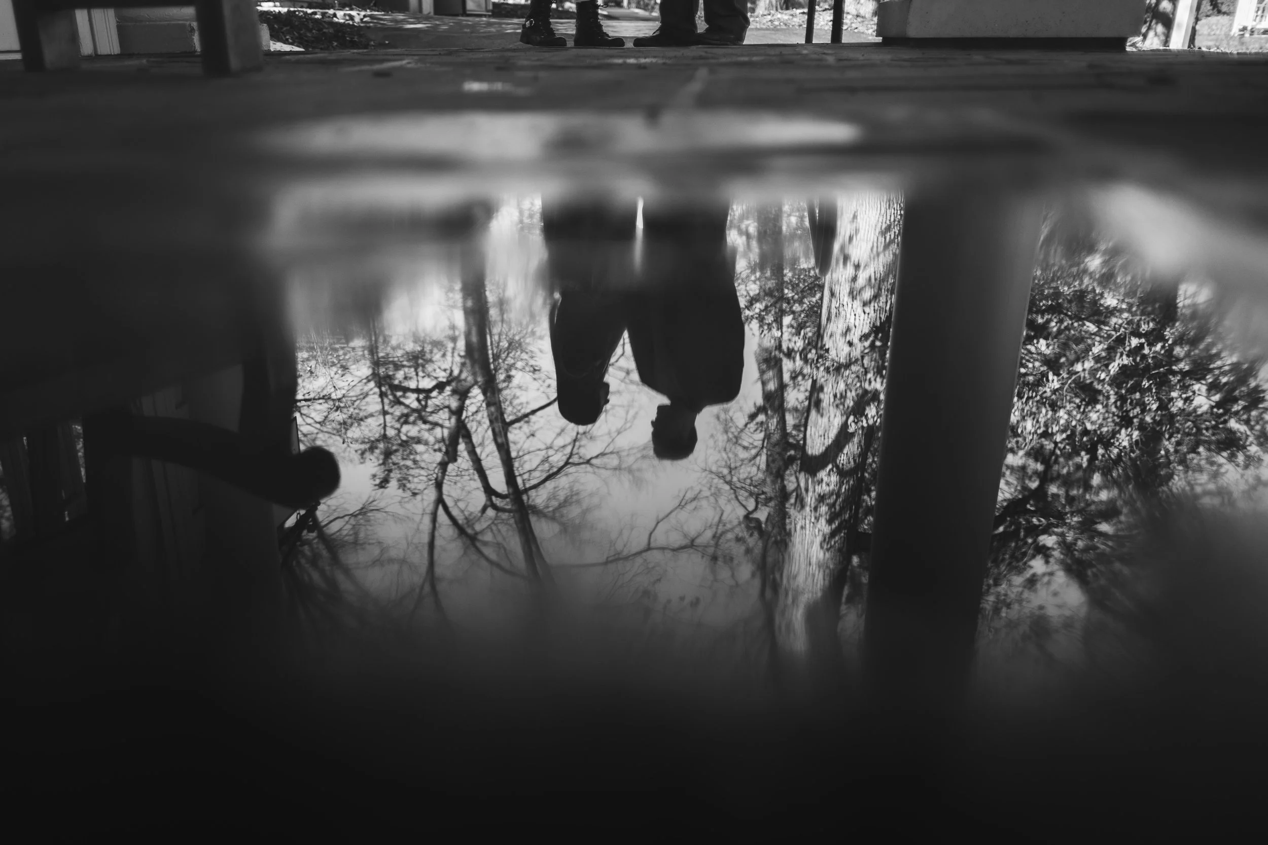 Reflection of two people standing outdoors near trees with bare branches, seen in a puddle or reflective surface, in black and white.