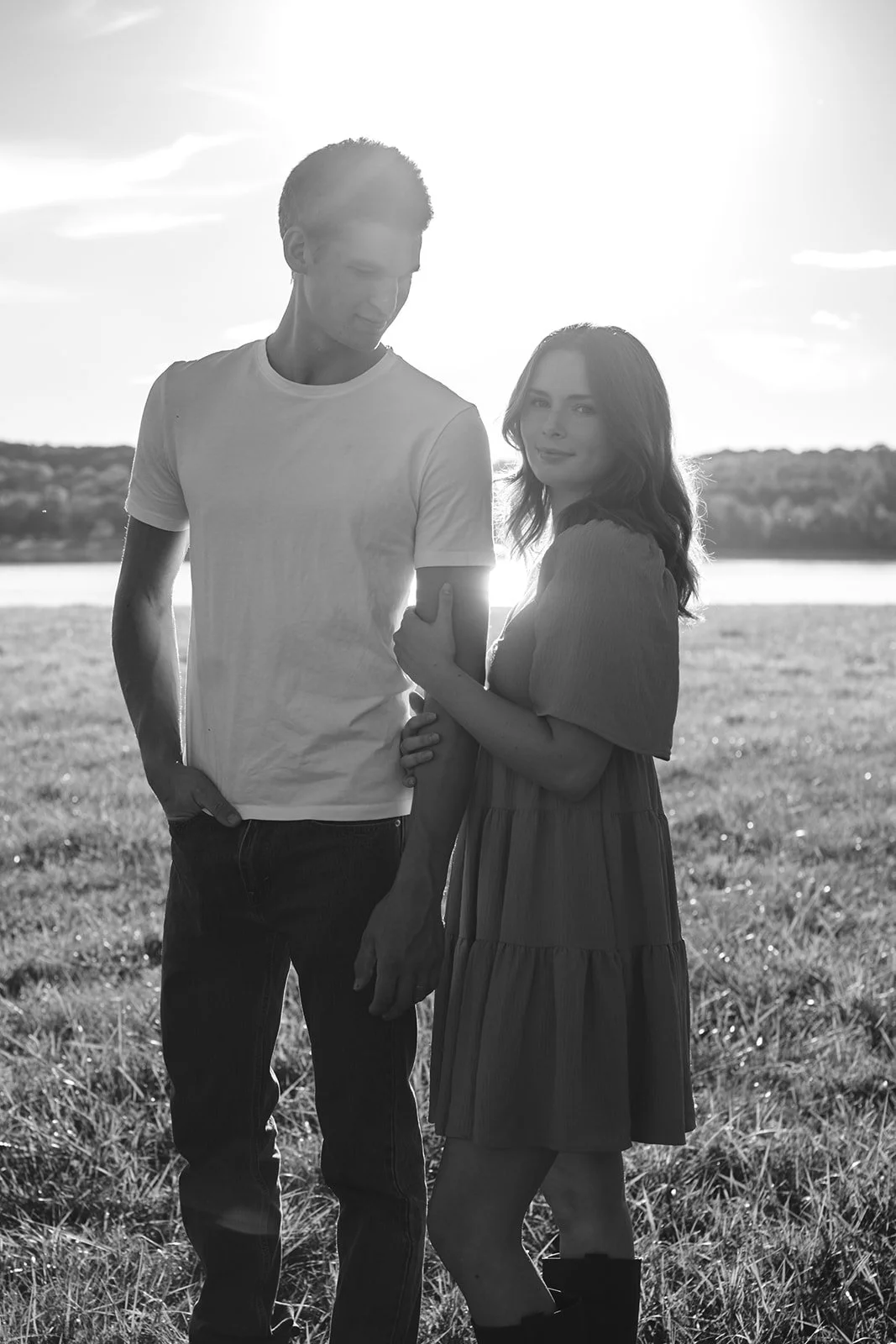 A black and white photo of a young man and woman standing outdoors on a grassy field near a body of water with trees in the background, backlit by the setting sun.