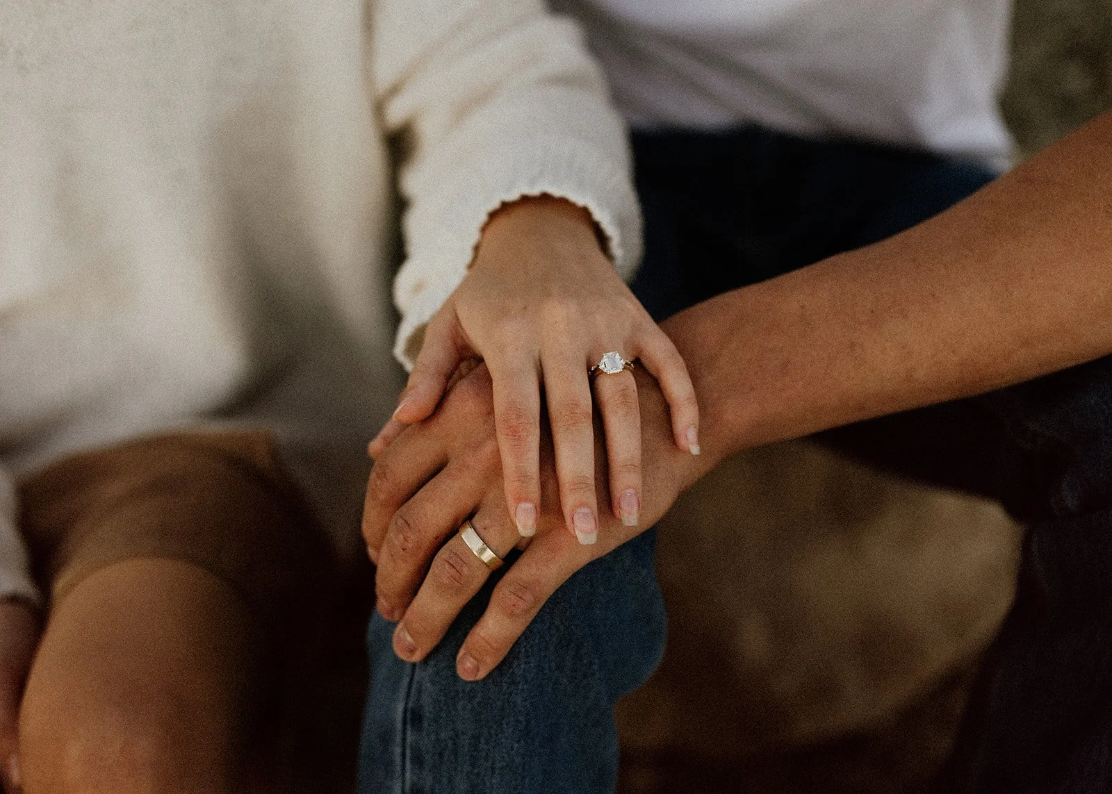 Close-up of a person wearing a wedding ring and an engagement ring, holding hands with another person. The person with rings is wearing a white long-sleeve shirt, and the other person has a tan skin tone, wearing a short-sleeve shirt. They are sittin