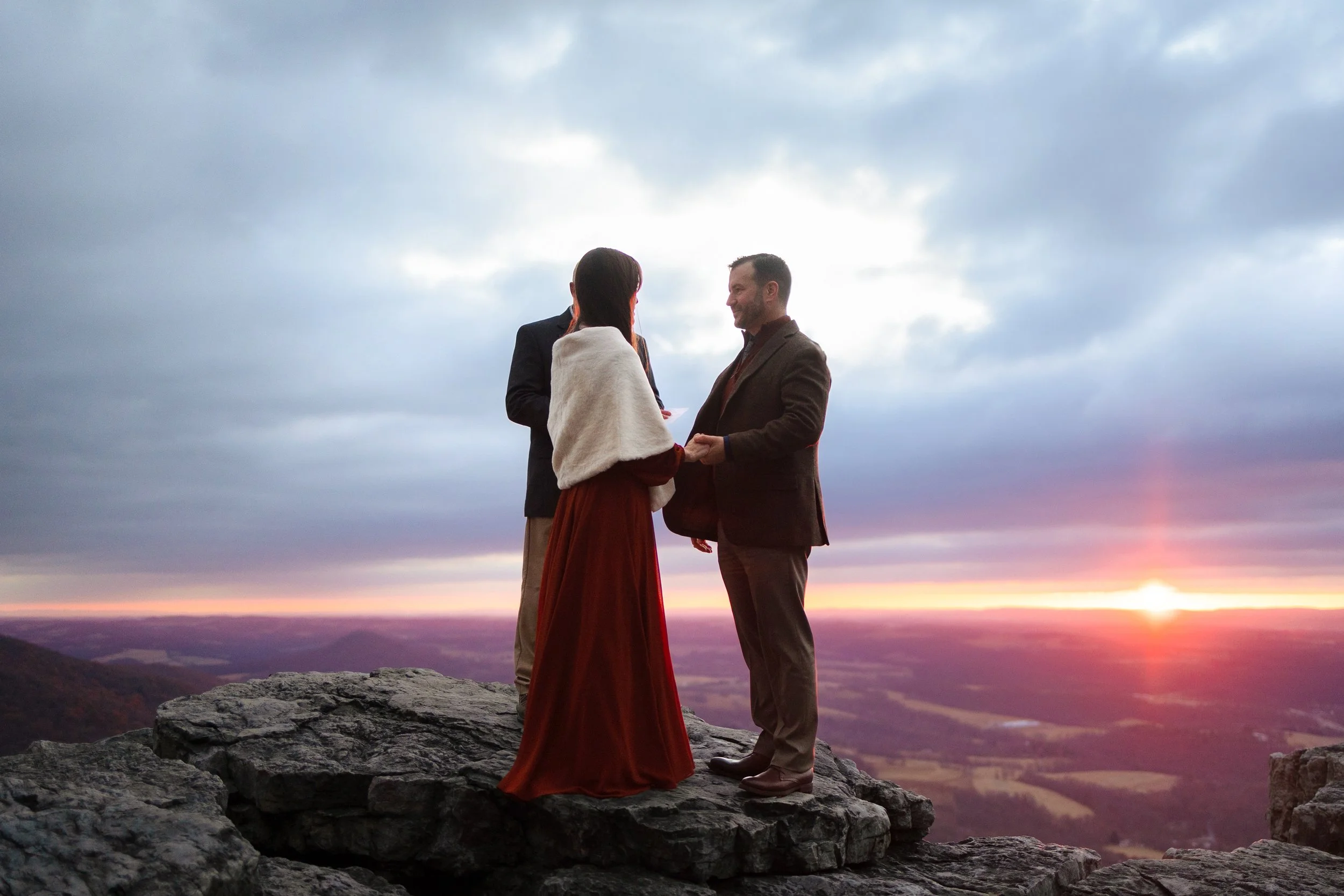 A couple stands on a rocky cliff at sunset, facing each other and holding hands, with a person officiating a ceremony behind them, overlooking a landscape with hills and a cloudy sky.