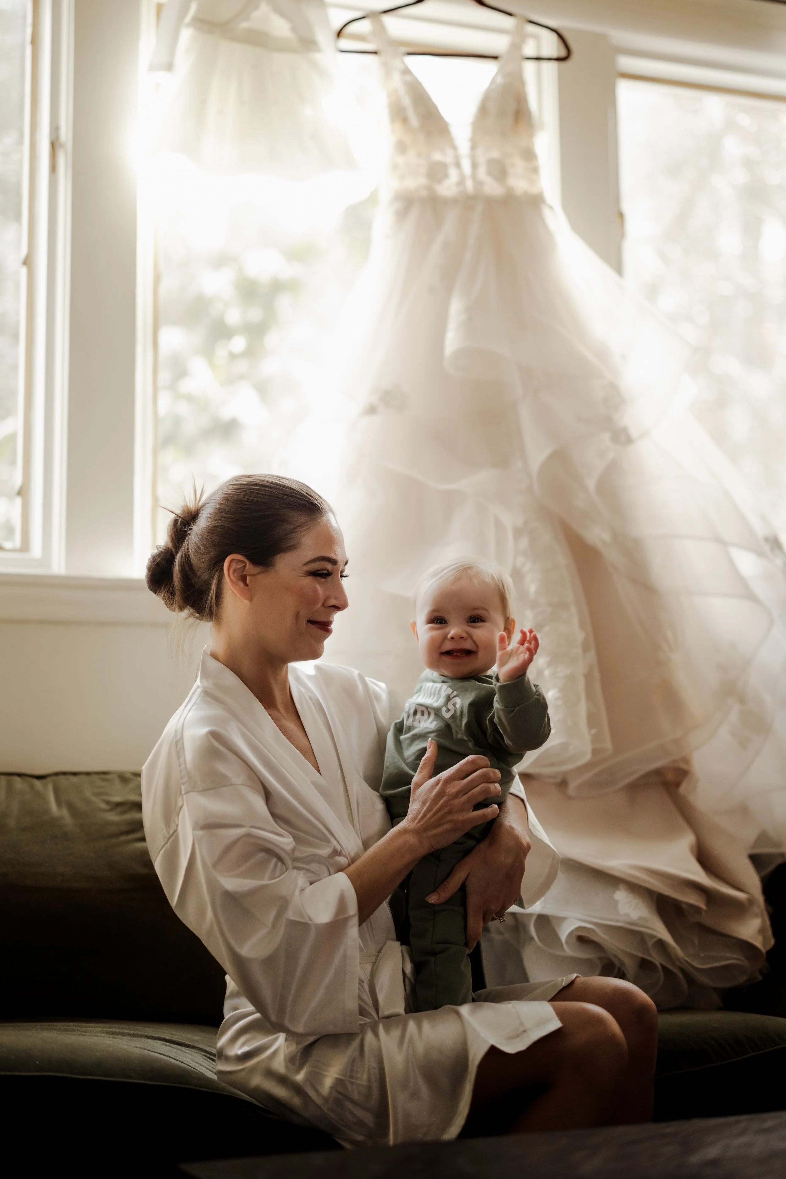 A woman sitting on a couch holding a smiling baby, with wedding dresses hanging in the background near a window.