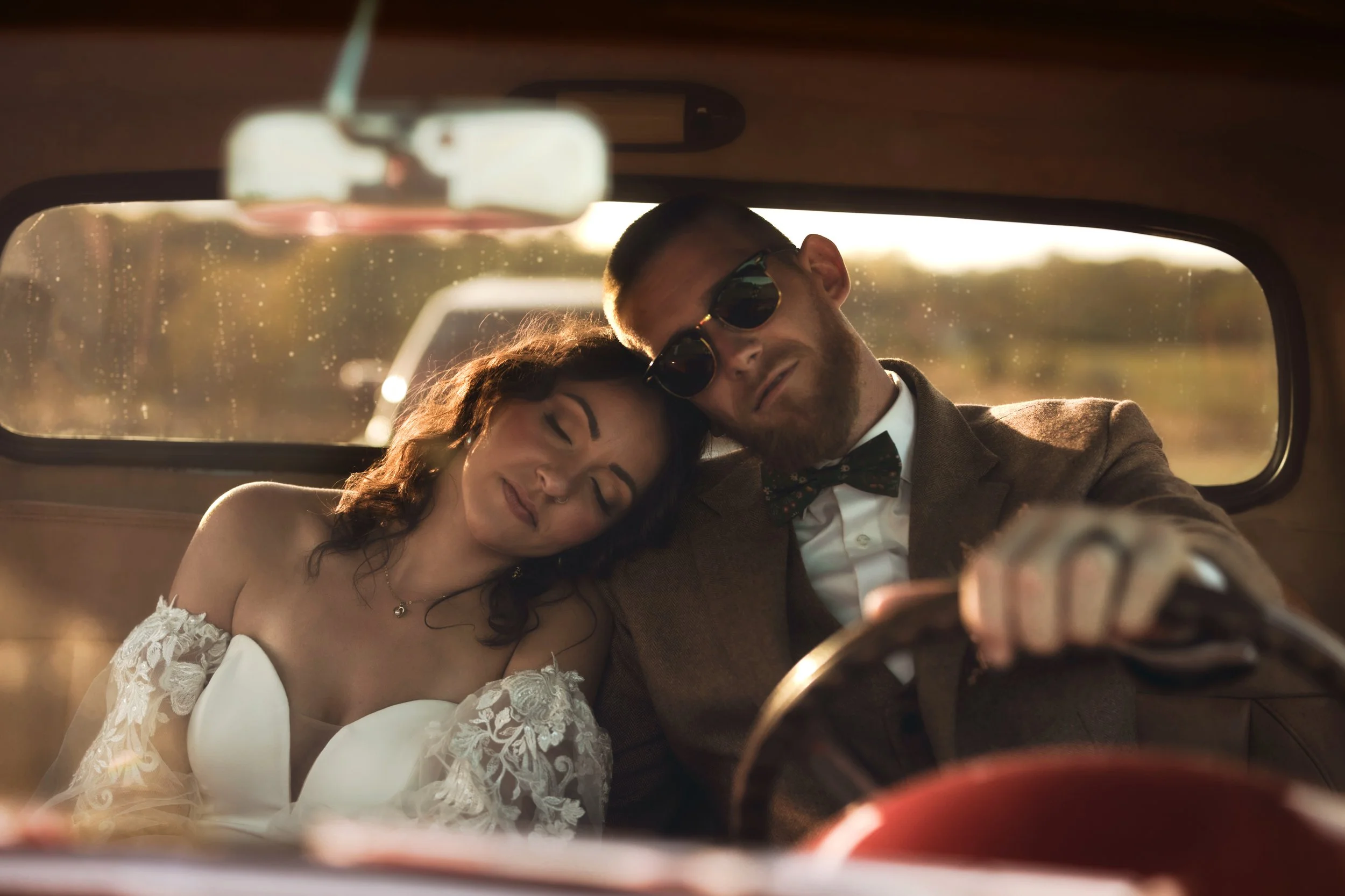 A man and a woman sitting inside a car, resting their heads close together. The woman has her eyes closed and is wearing a white off-shoulder dress with lace details, while the man is wearing sunglasses, a brown suit, and a bowtie, looking at the cam