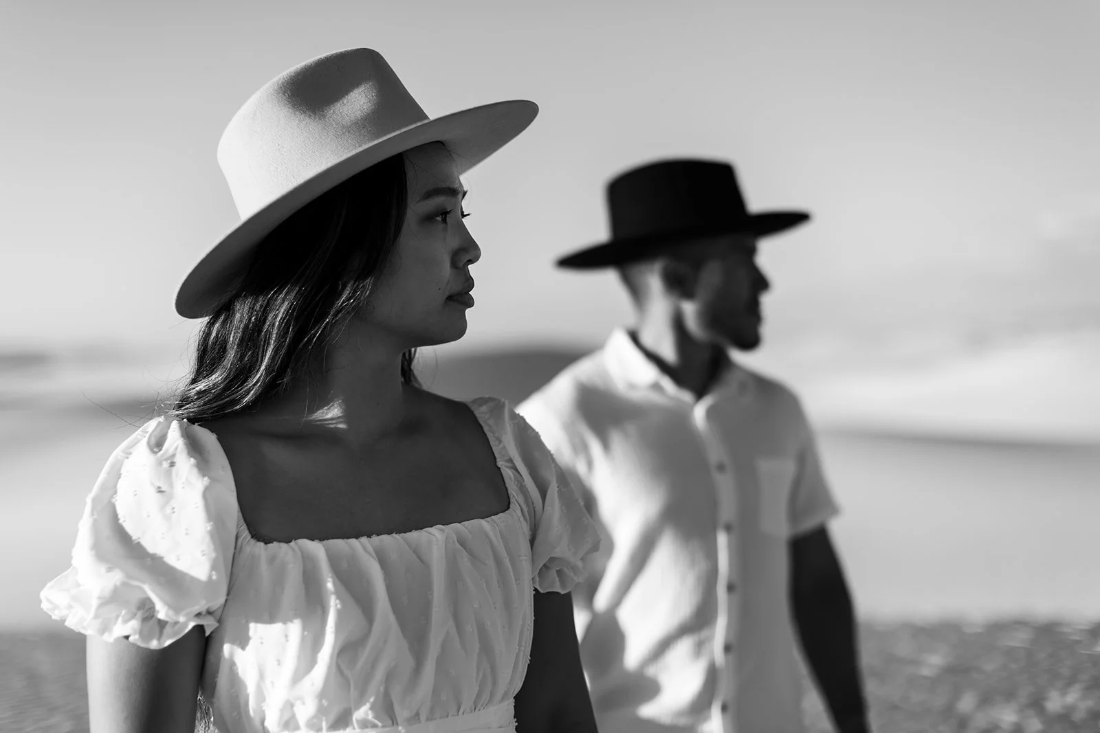 Black and white photo of a woman and a man walking on a beach, both wearing wide-brimmed hats and casual clothing, facing sideways.