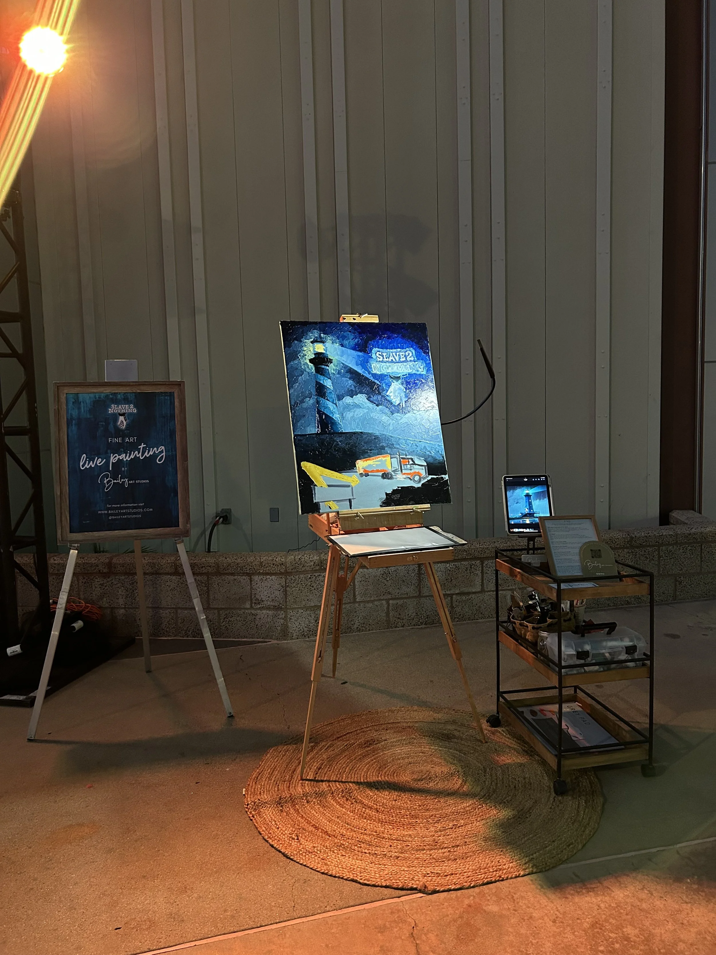 An art display setup featuring a painting of a lighthouse at night with a blue sky, waves, and a vehicle, along with a sign indicating live painting at Bailey Art Studios. The scene includes a cylindrical woven rug on the floor, a small wheeled cart with art supplies, a framed poster on an easel, and a laptop displaying the artwork, all in an indoor space with a wood-paneled wall.