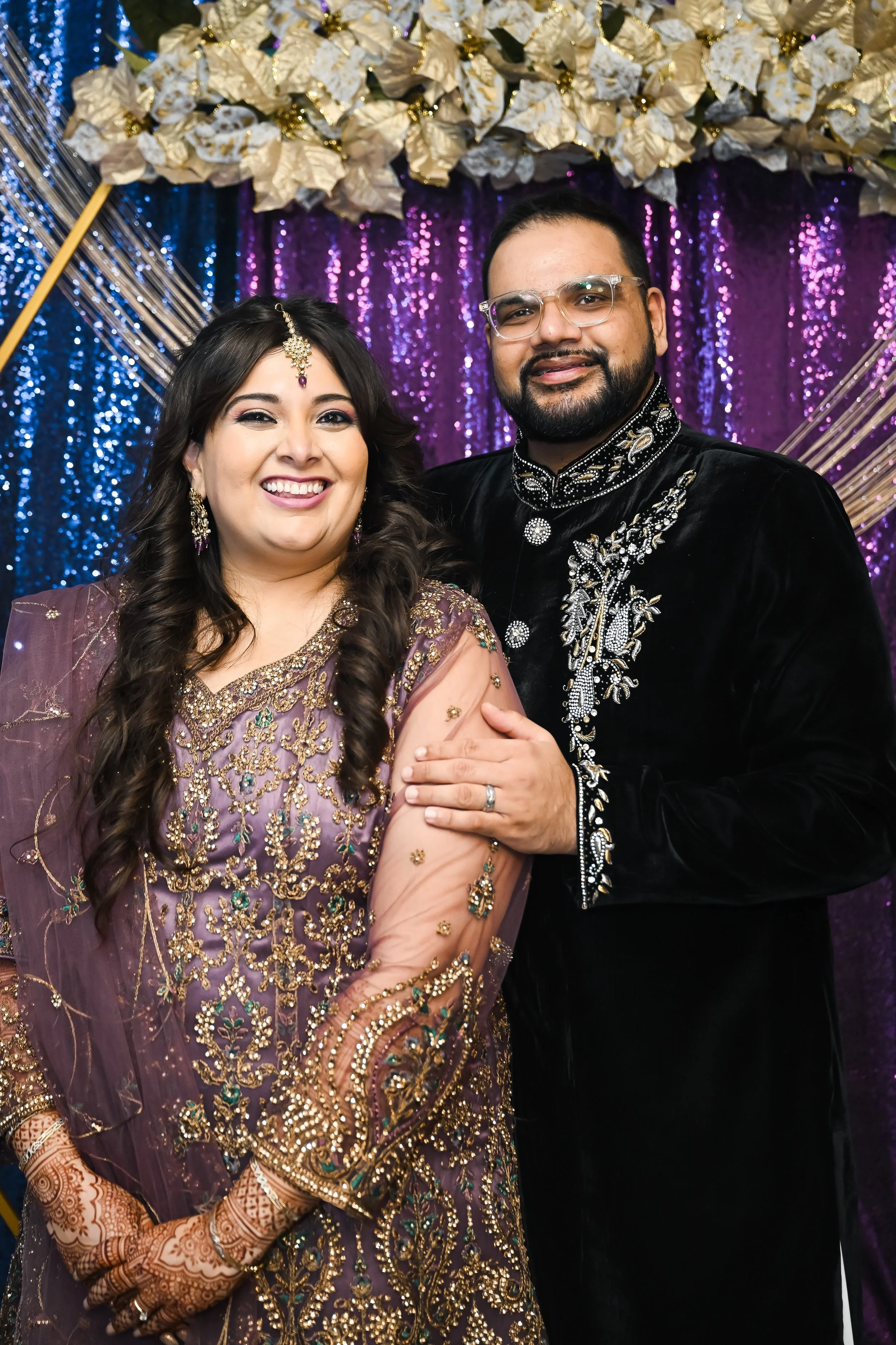 A smiling couple dressed in traditional Indian wedding attire, standing in front of a colorful backdrop with flowers and purple sequined fabric.