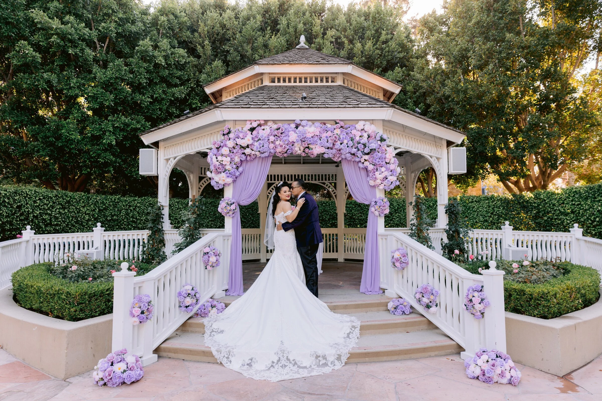 A bride and groom standing in a gazebo decorated with purple and pink flowers during a wedding ceremony.